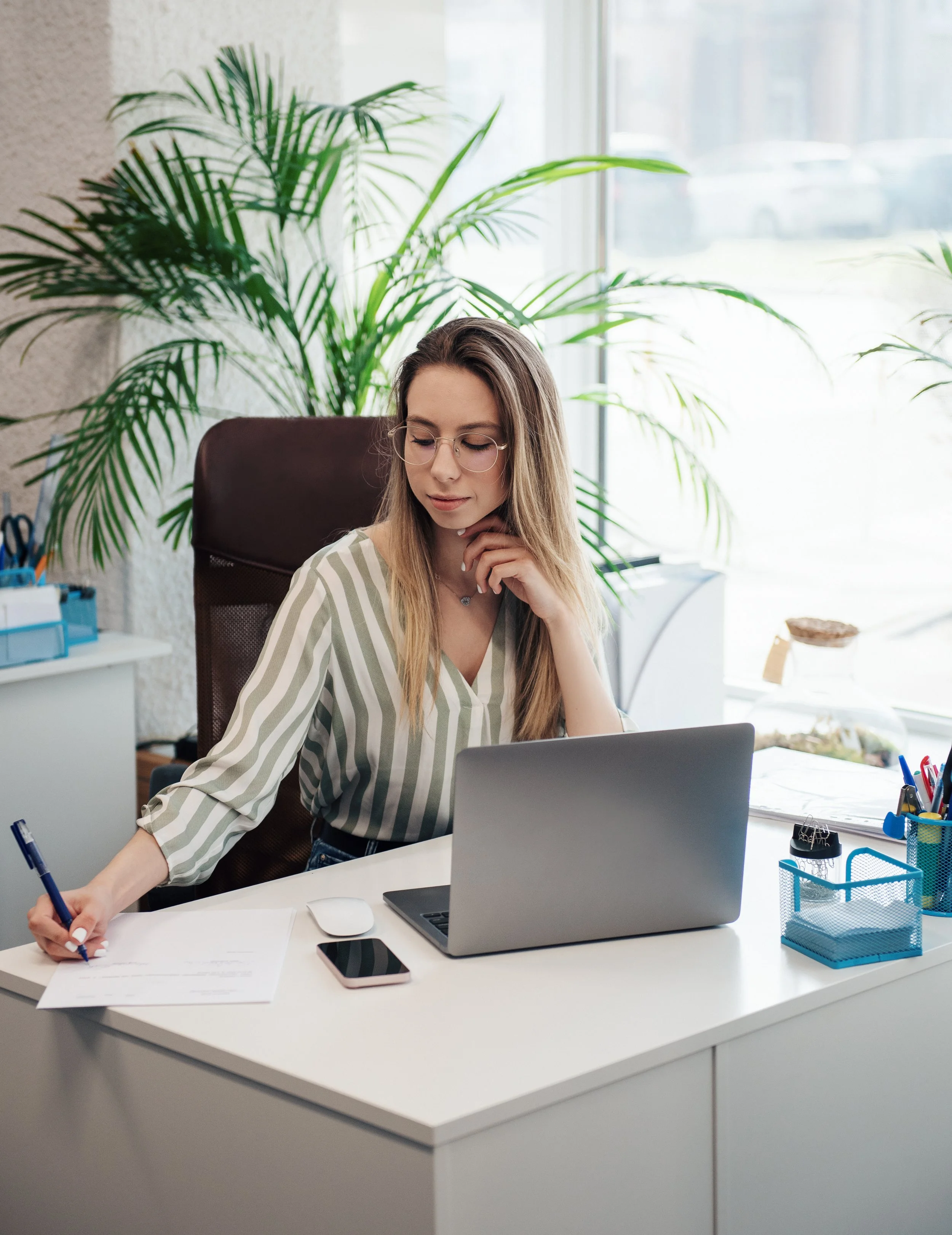 A young woman with glasses and long hair working at a white desk in an office, surrounded by office supplies and a large green plant.