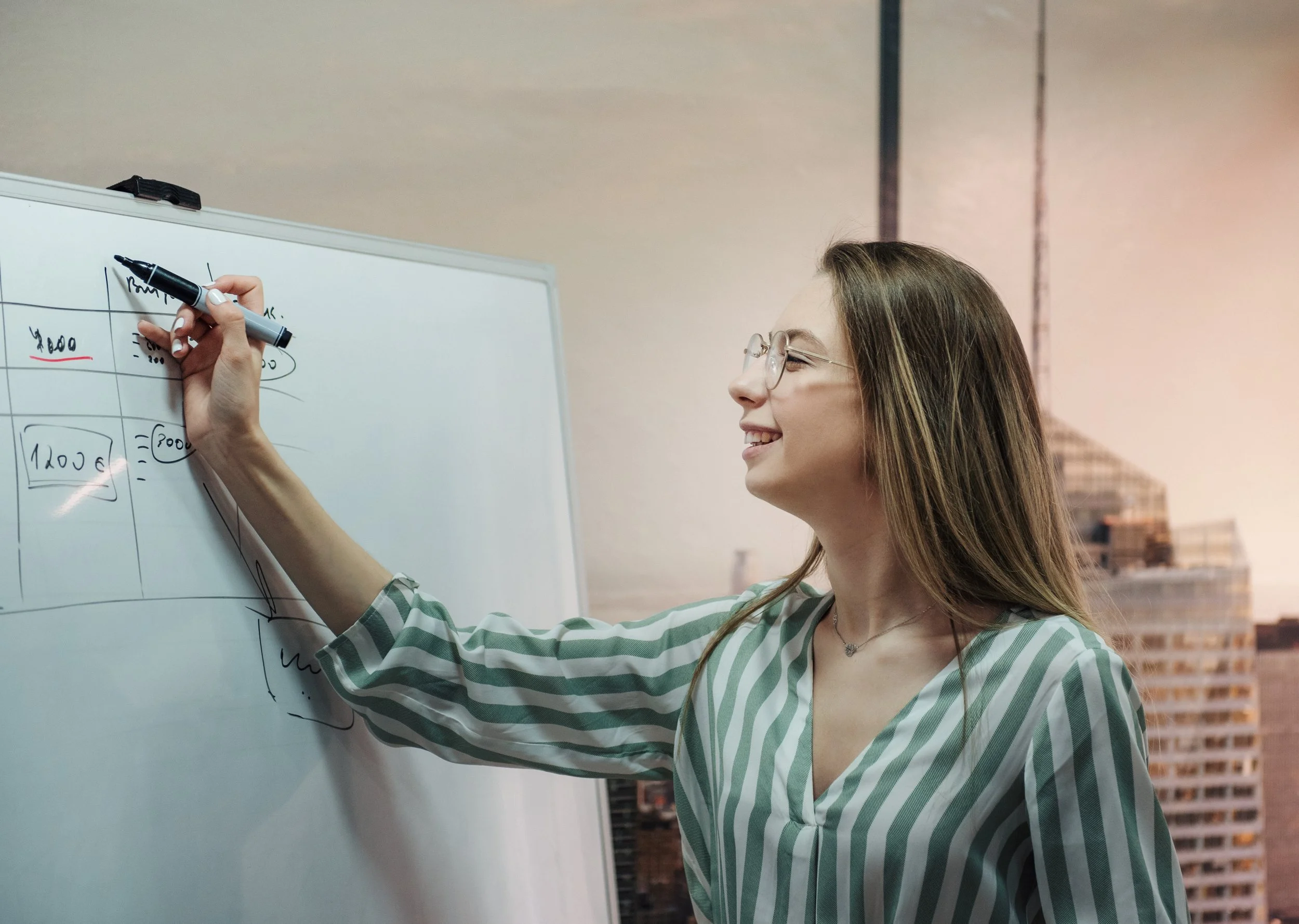 A smiling woman wearing glasses and a green and white striped shirt writes on a whiteboard in a room with a brick wall and shelves in the background.