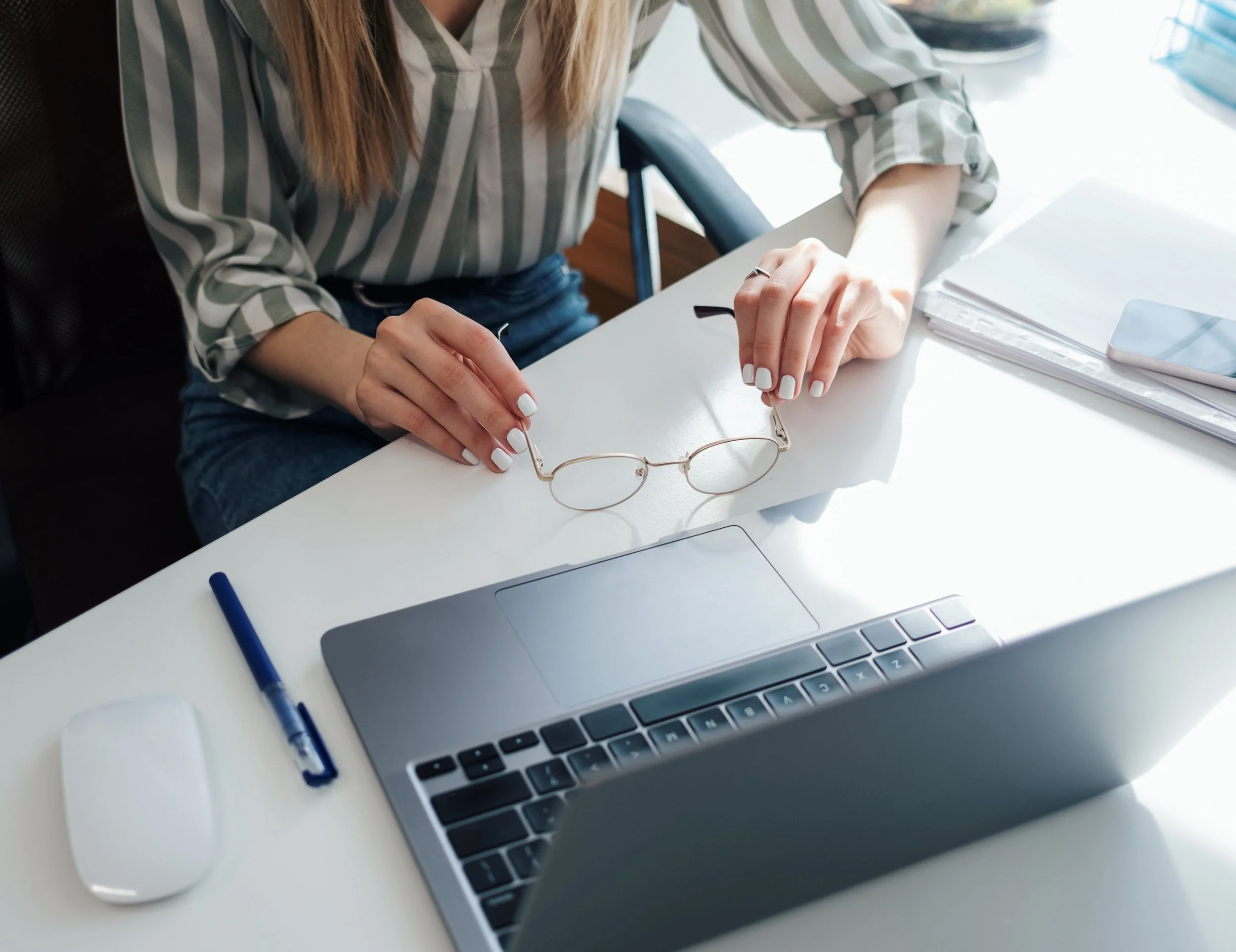 Close-up of a woman sitting at a desk with a laptop, eyeglasses, and a blue pen. She is wearing a striped shirt, and there is a white wireless device and a partially visible notebook on the desk.