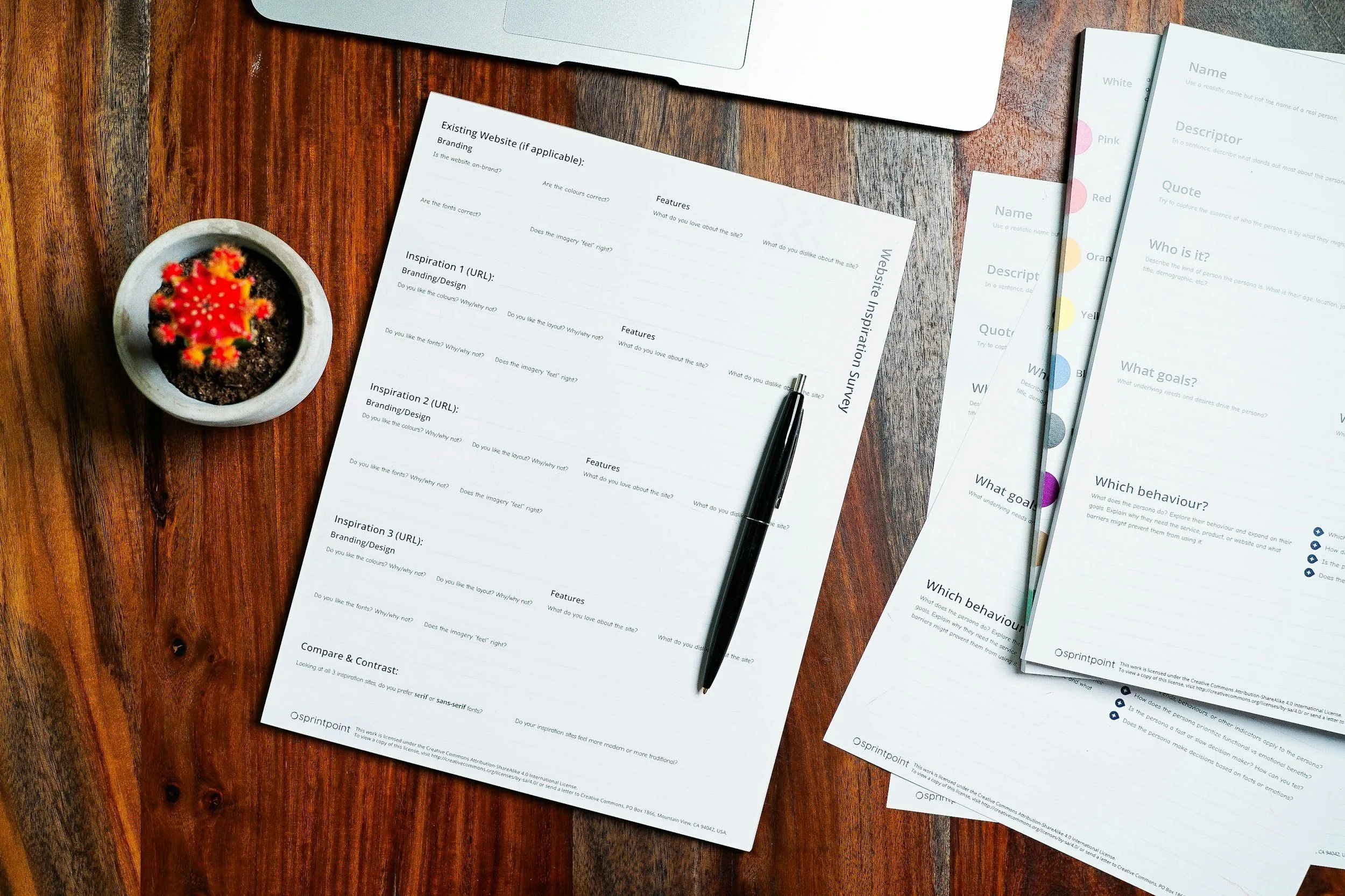 A wooden desk with printed survey forms, a black pen, a small potted cactus, and some folders or sheets of paper on top.