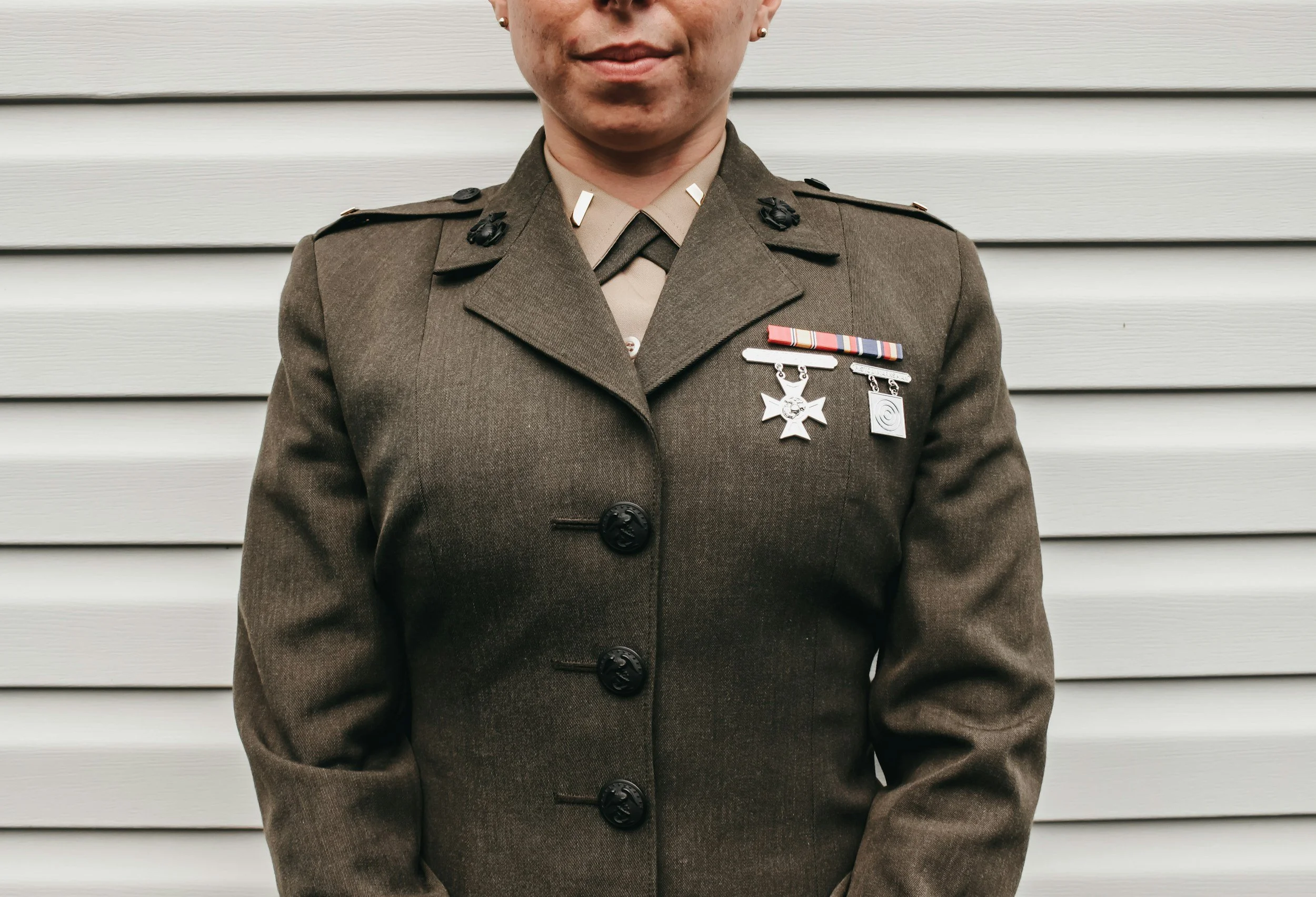 A person in a military uniform standing outdoors in front of light-colored horizontal siding. The person is wearing medals and insignia on their uniform.