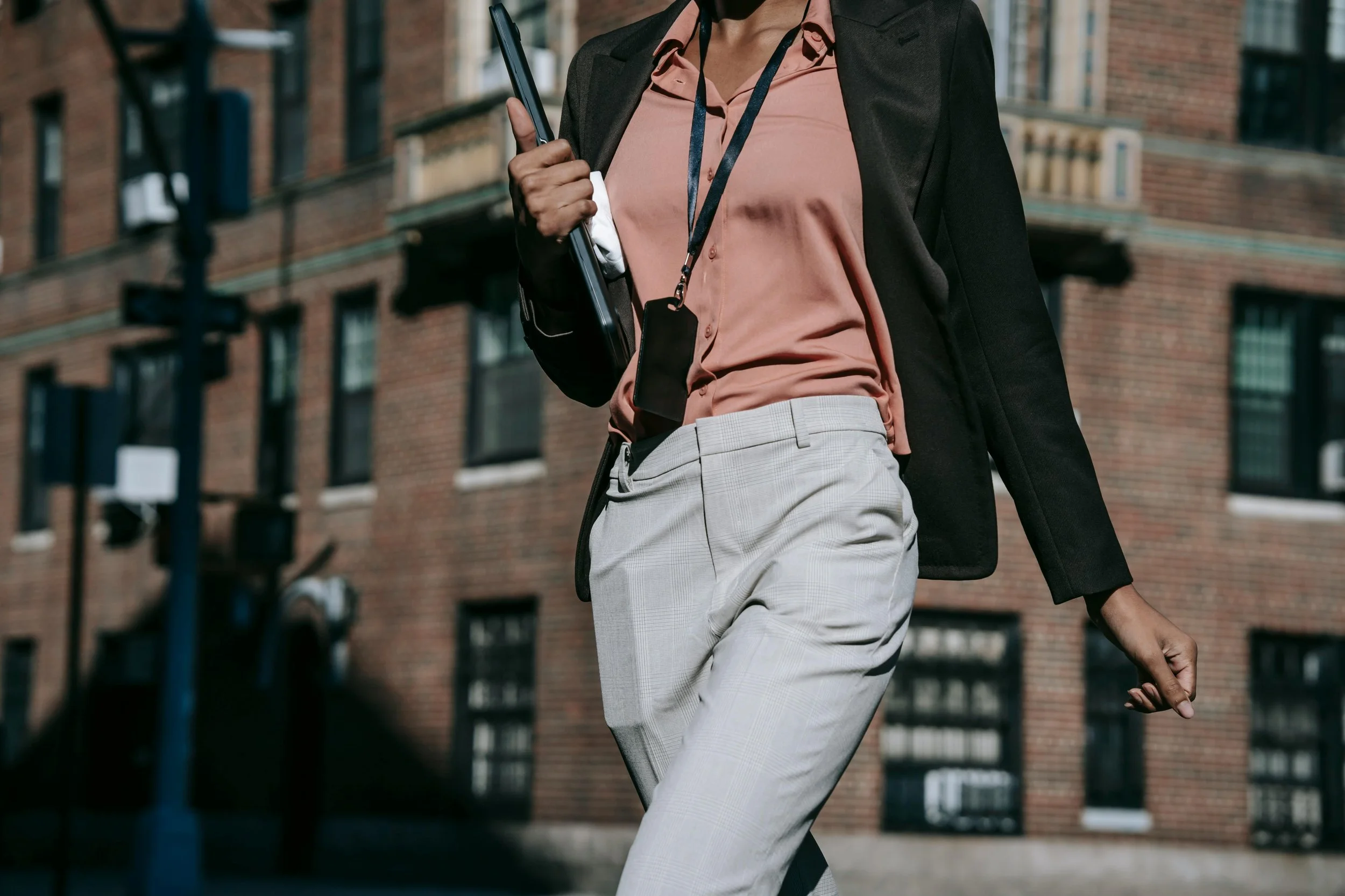 A woman in business attire carrying a backpack and holding a tablet, walking outdoors in an urban setting with brick apartment buildings in the background.