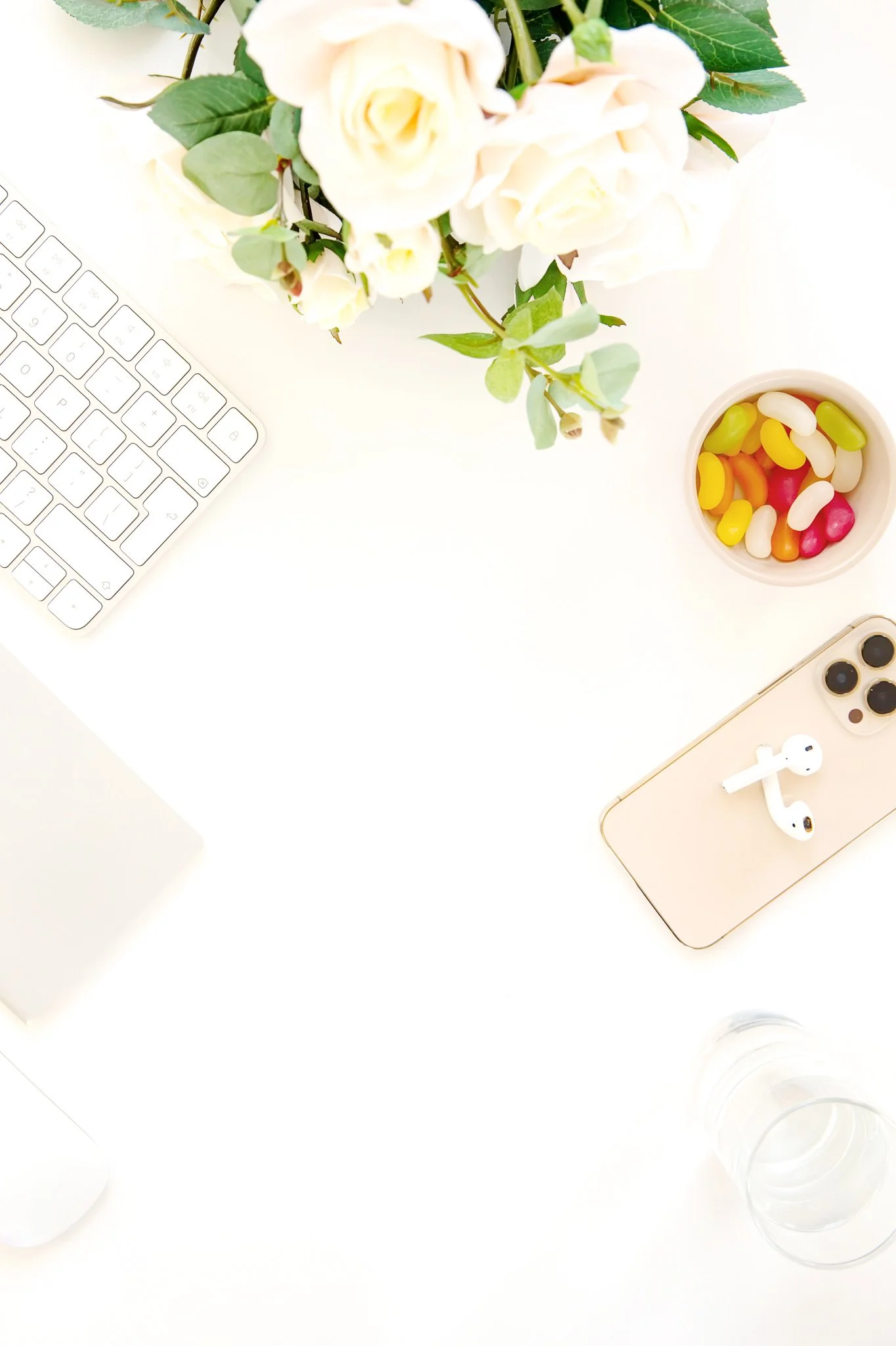 A white workspace desk with a computer keyboard, a vase of white roses and greenery, a bowl of colorful jelly beans, a smartphone with wireless earbuds, and several empty glasses.