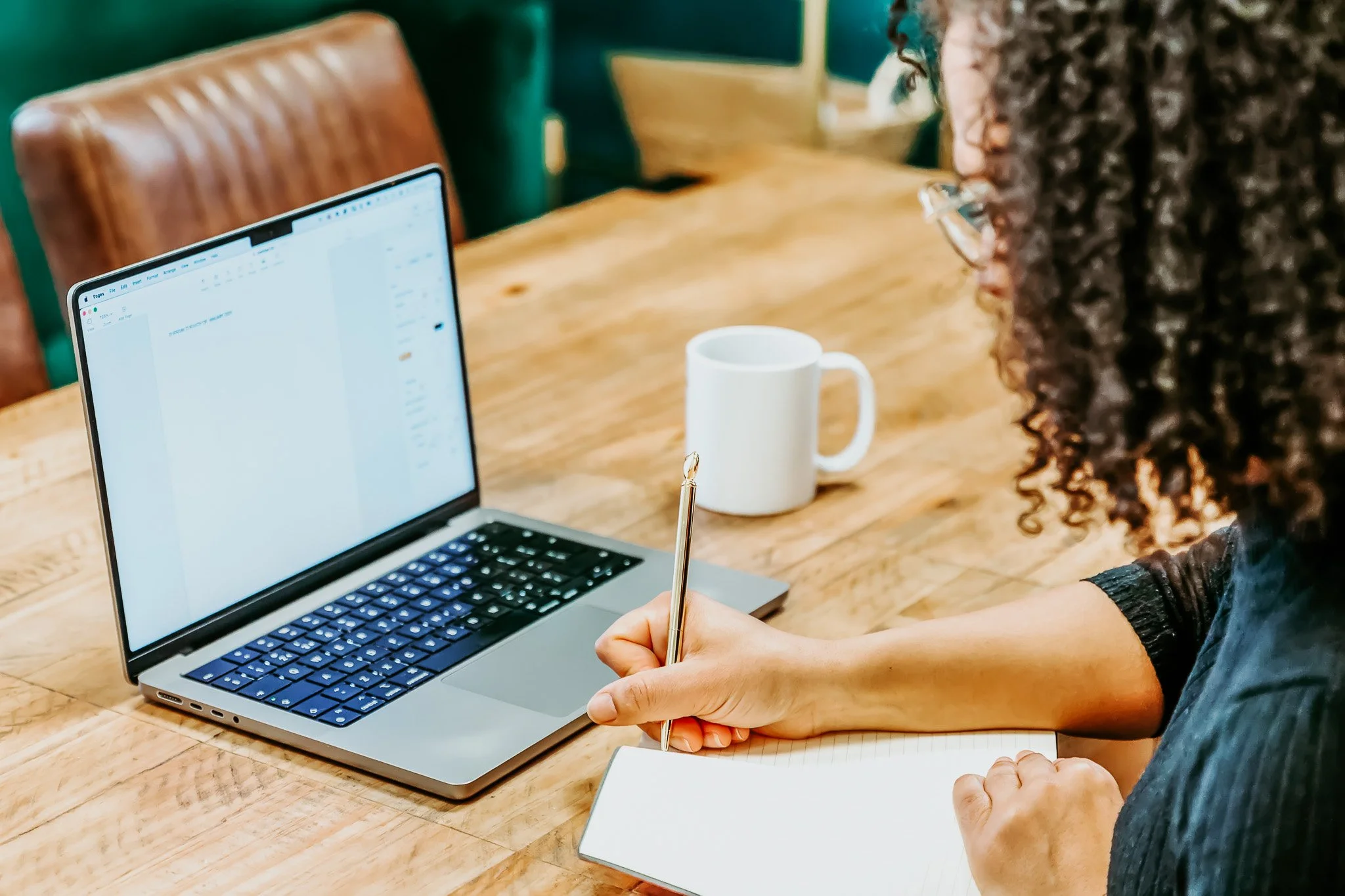 A woman with curly hair sitting at a wooden table, working on a laptop, taking notes in a notebook with a pen, with a white coffee mug nearby.