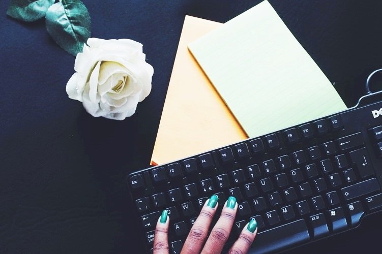 Close-up of a person's hand with green nail polish typing on a black computer keyboard, with a white rose, green leaves, and two notebooks, one yellow and one white, on a dark surface.