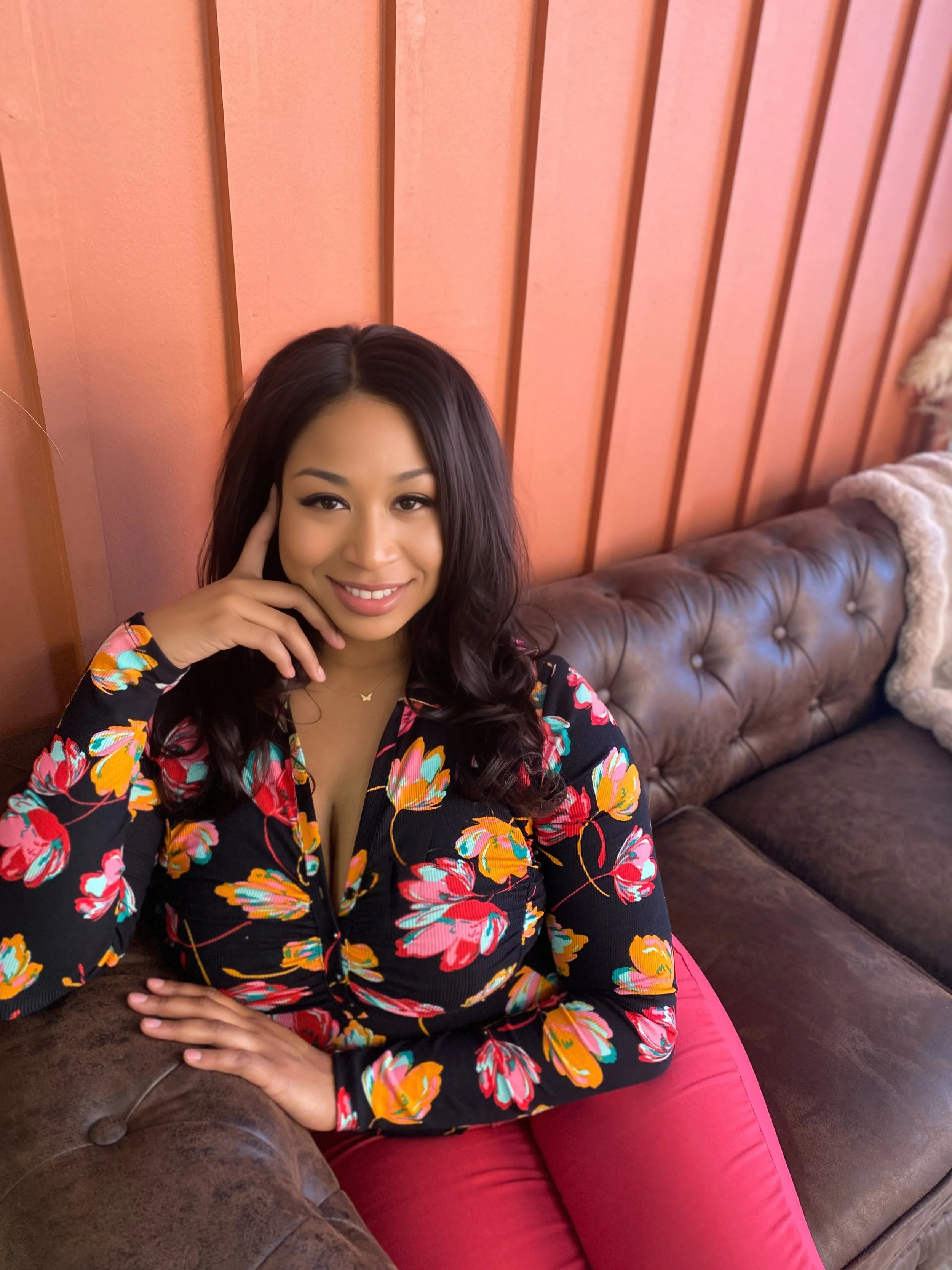 A woman sitting on a brown leather couch, smiling, with her left hand holding her face and her right arm resting on the couch. She is wearing a black floral top with pink, orange, and yellow flowers, and pink pants. The background features a pink wooden-paneled wall.