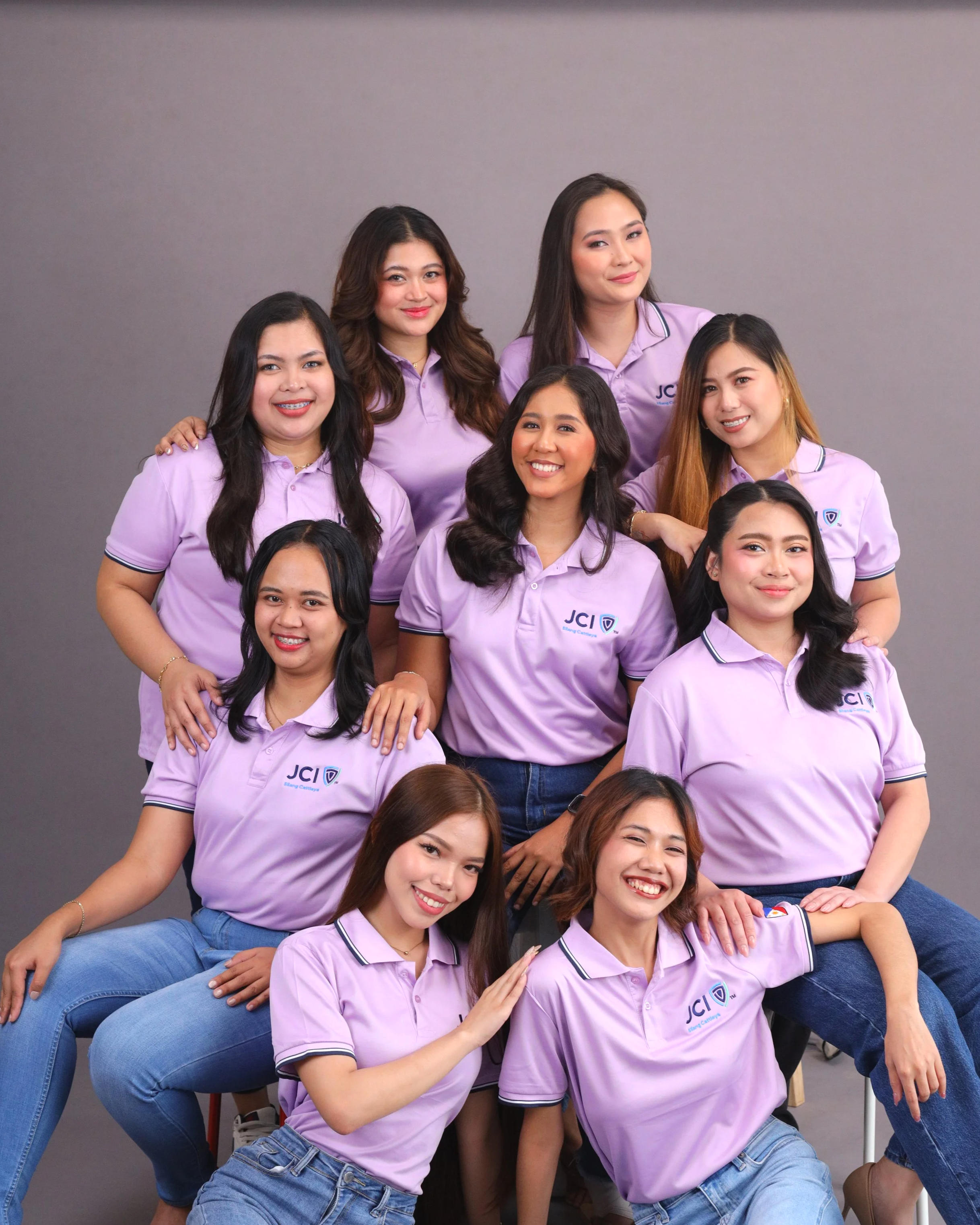 Group of young women wearing matching lavender polo shirts with 'JCI' logo, posing for a photo against a plain grey background, smiling and showing camaraderie.