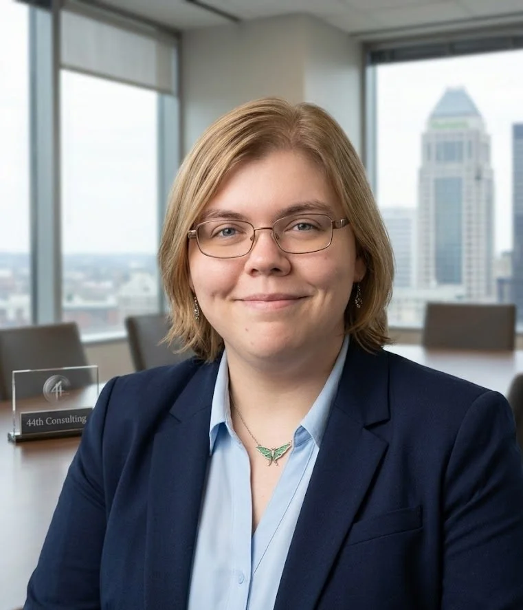 Businesswoman in a navy blazer and glasses sitting in a conference room with large windows and city skyline view.