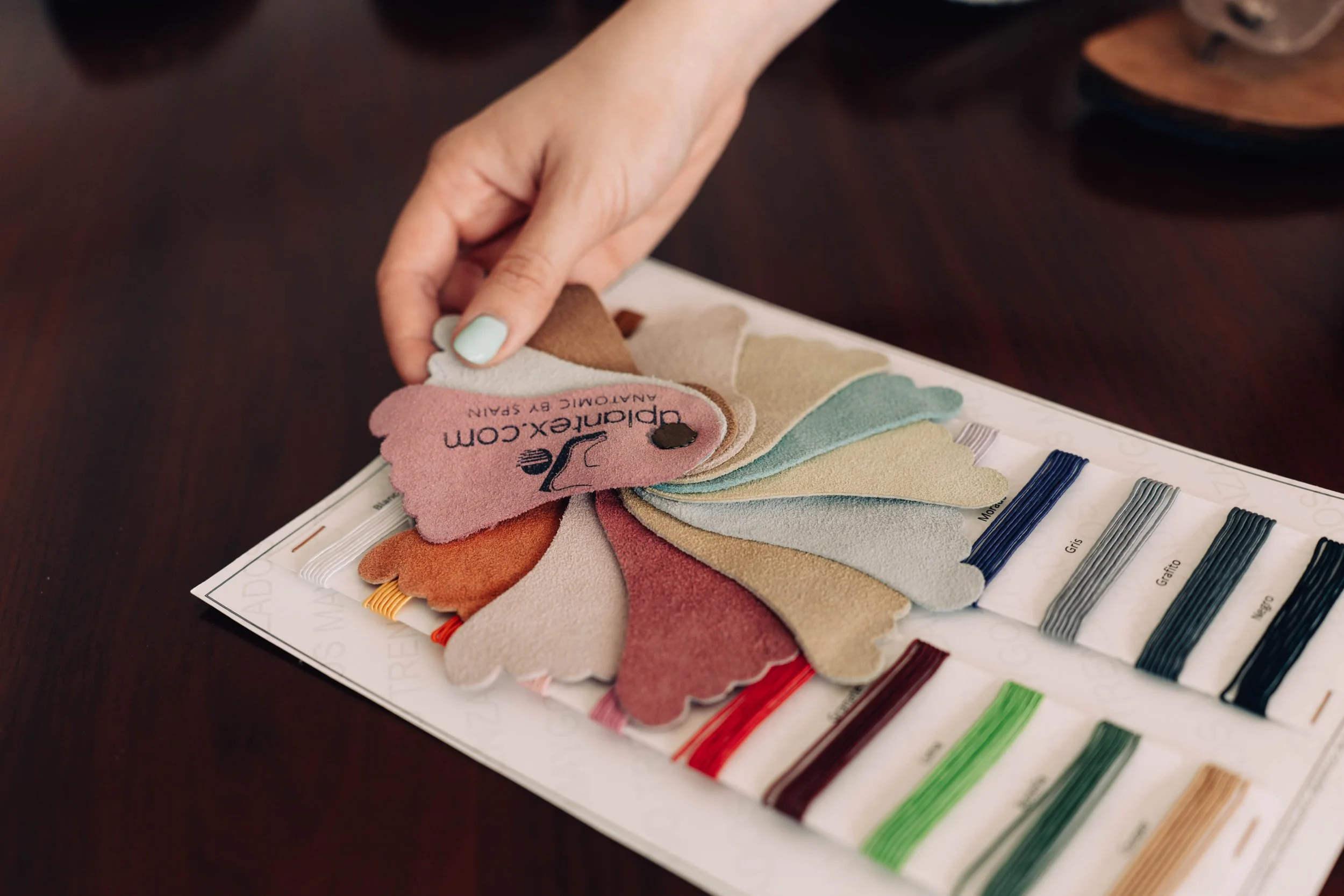 Muestra en una tarjeta de muestras de colores y texturas de telas, con una mano señalando las muestras. Incluye etiquetas con nombres de colores como gris, granate, entre otros.