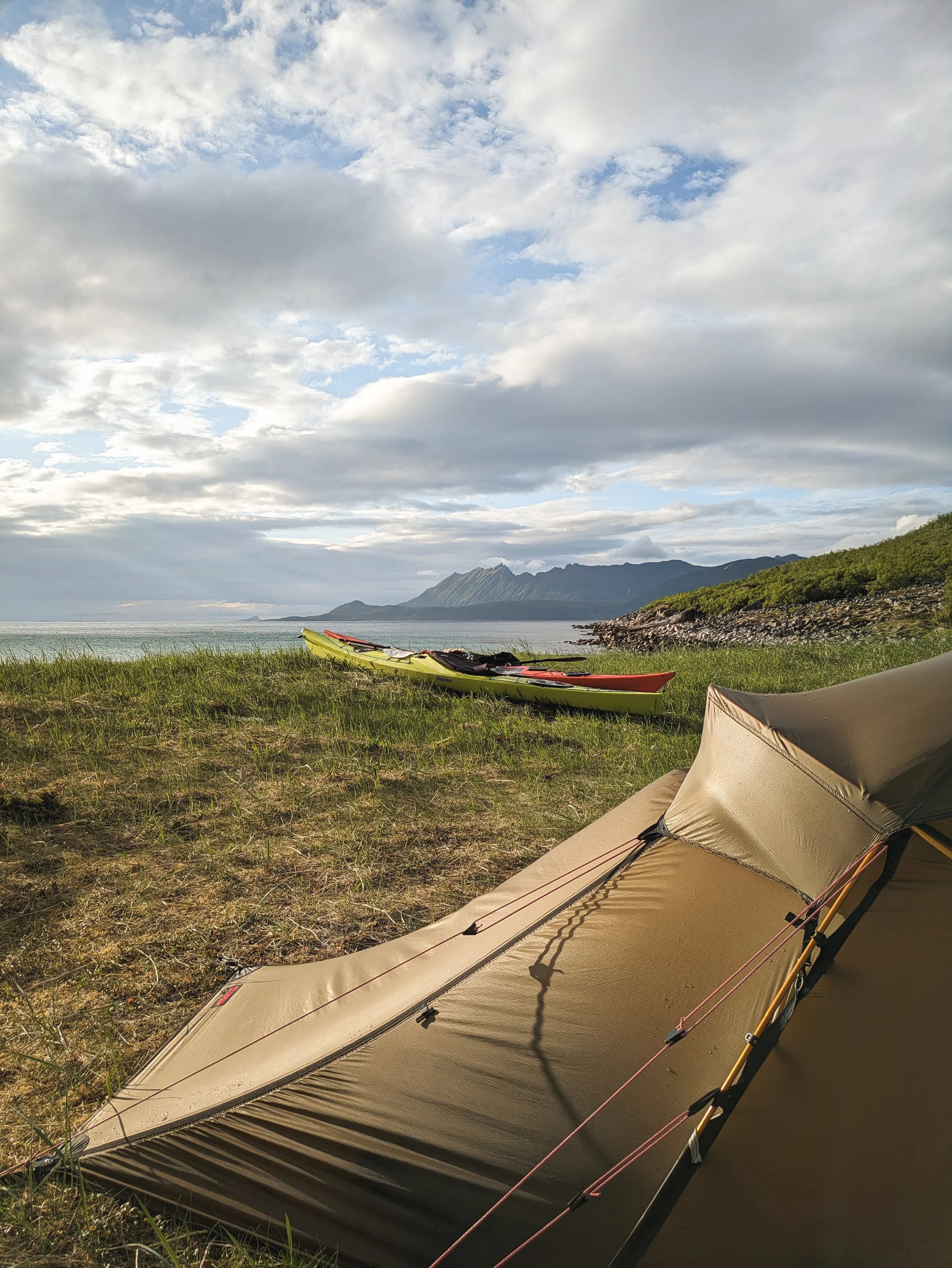 Camping spot on a remote coastline with tent and kayaks in the foreground and sea and mountains in the background