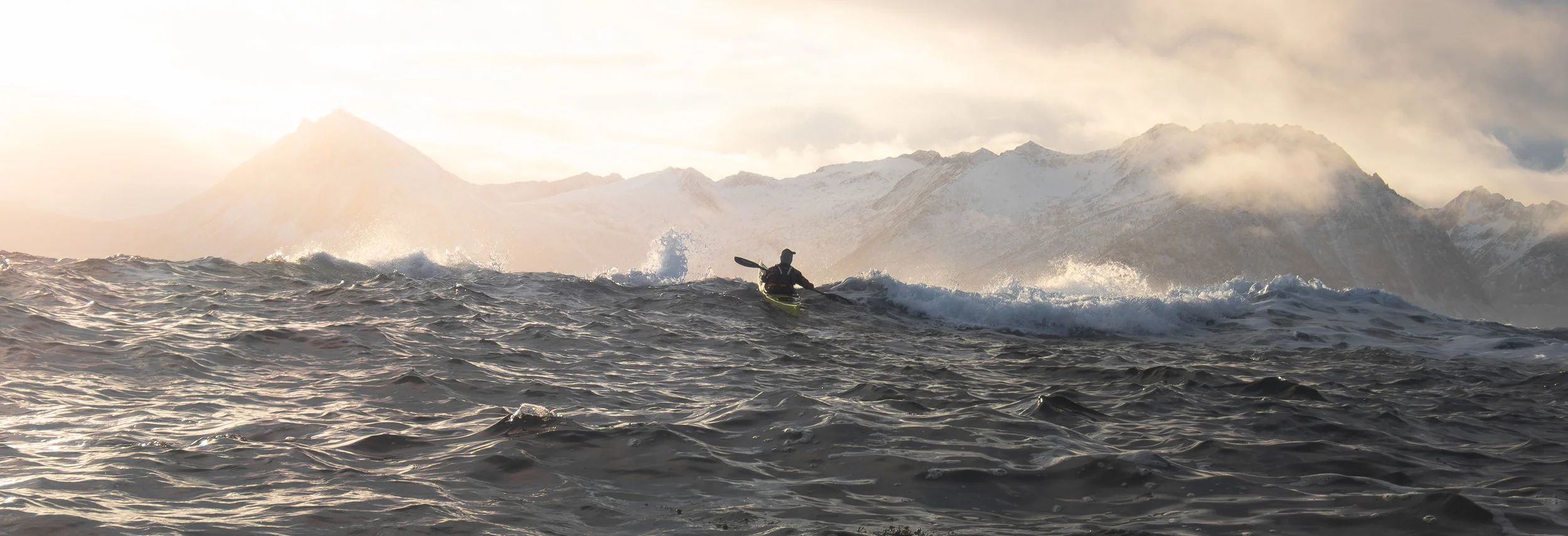 Lone kayaker on rough sea with dramatic mountain backdrop bathed in orange light