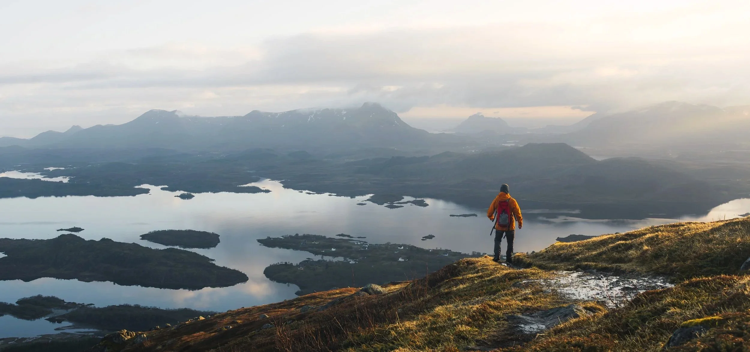 Man walking across a mountain with low sidelight and a backdrop of mountains shrouded in cloud with a fjord below