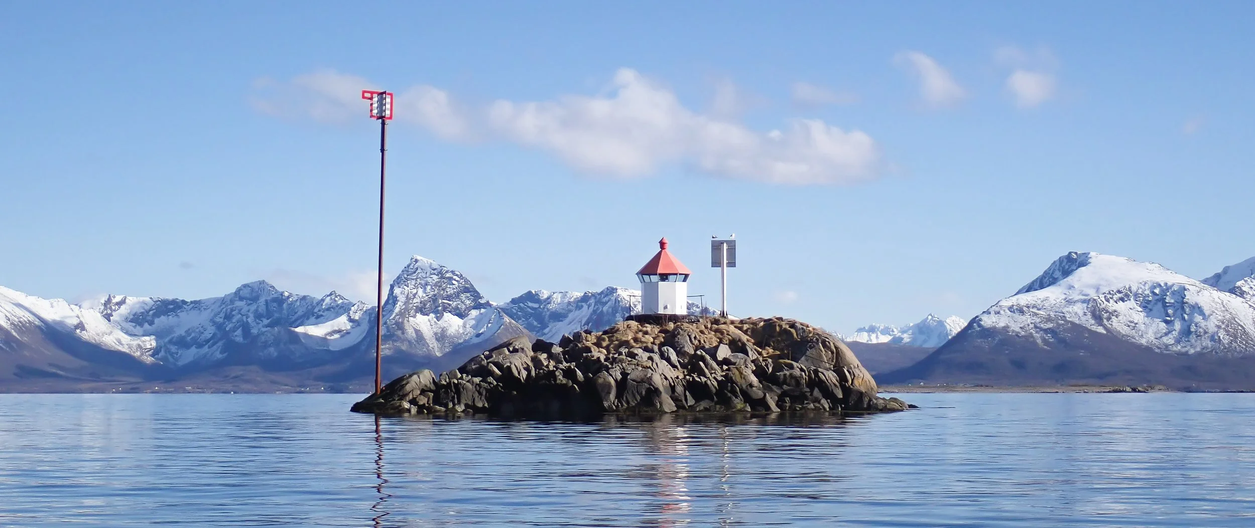 lighthouse on a small island with a backdrop of snow topped mountains in bright spring sunshine