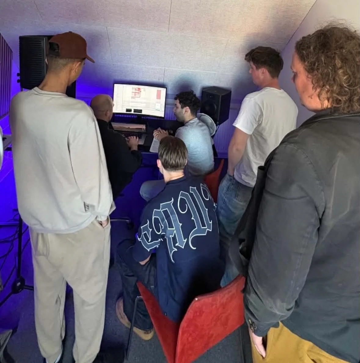 En gruppe mennesker i et musikstudie, hvor nogle arbejder på en computer med stor skærm, og andre står omkring og observerer.