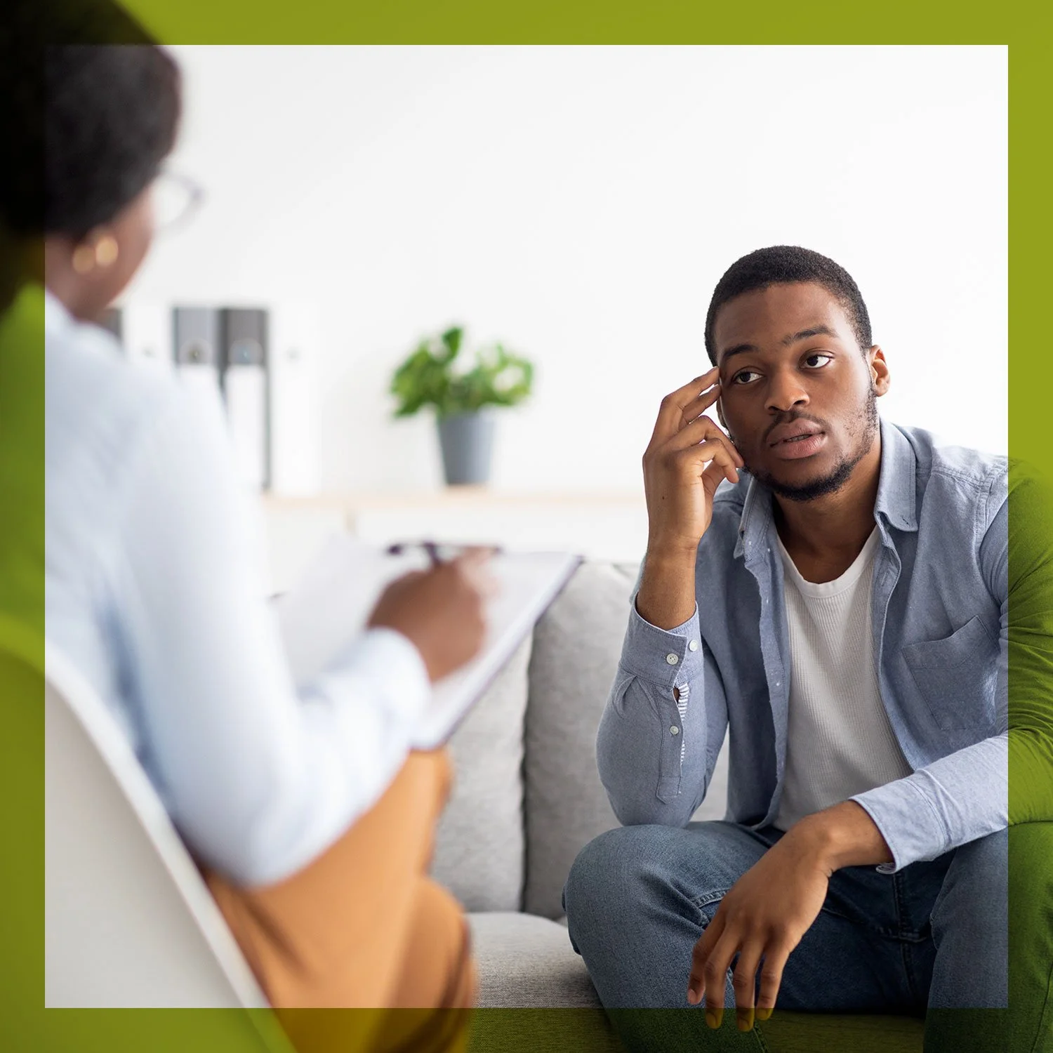 Young man in a light gray shirt sitting on a sofa during a therapy session, listening to a therapist holding a clipboard, with a potted plant in the background.