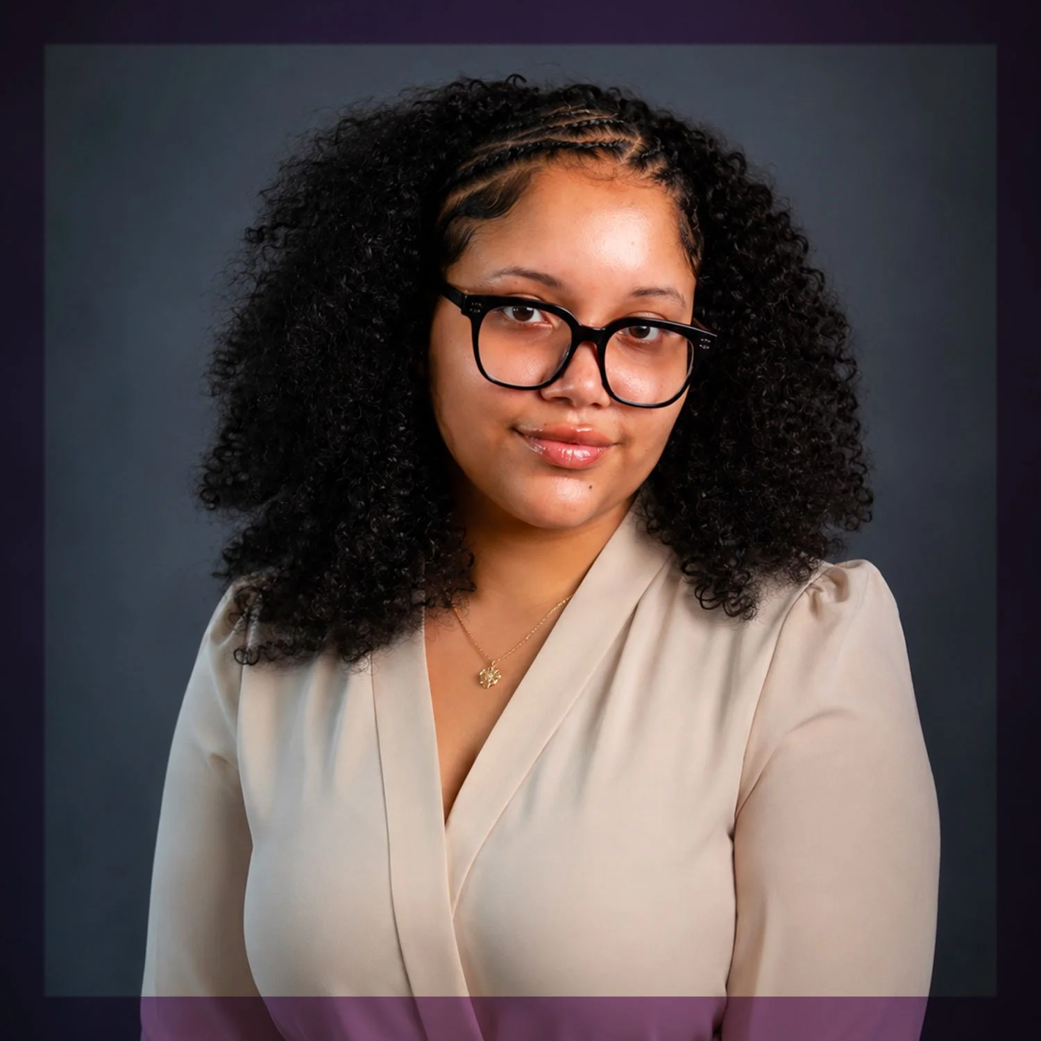 A woman with curly black hair, wearing black glasses, a beige blouse, and a gold necklace, posing against a dark background.