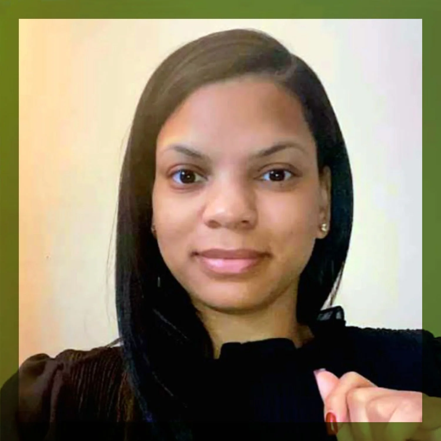 A young woman with straight black hair and light brown skin looking at the camera with a neutral expression. She is wearing a dark top and earrings, and is standing against a light-colored wall.