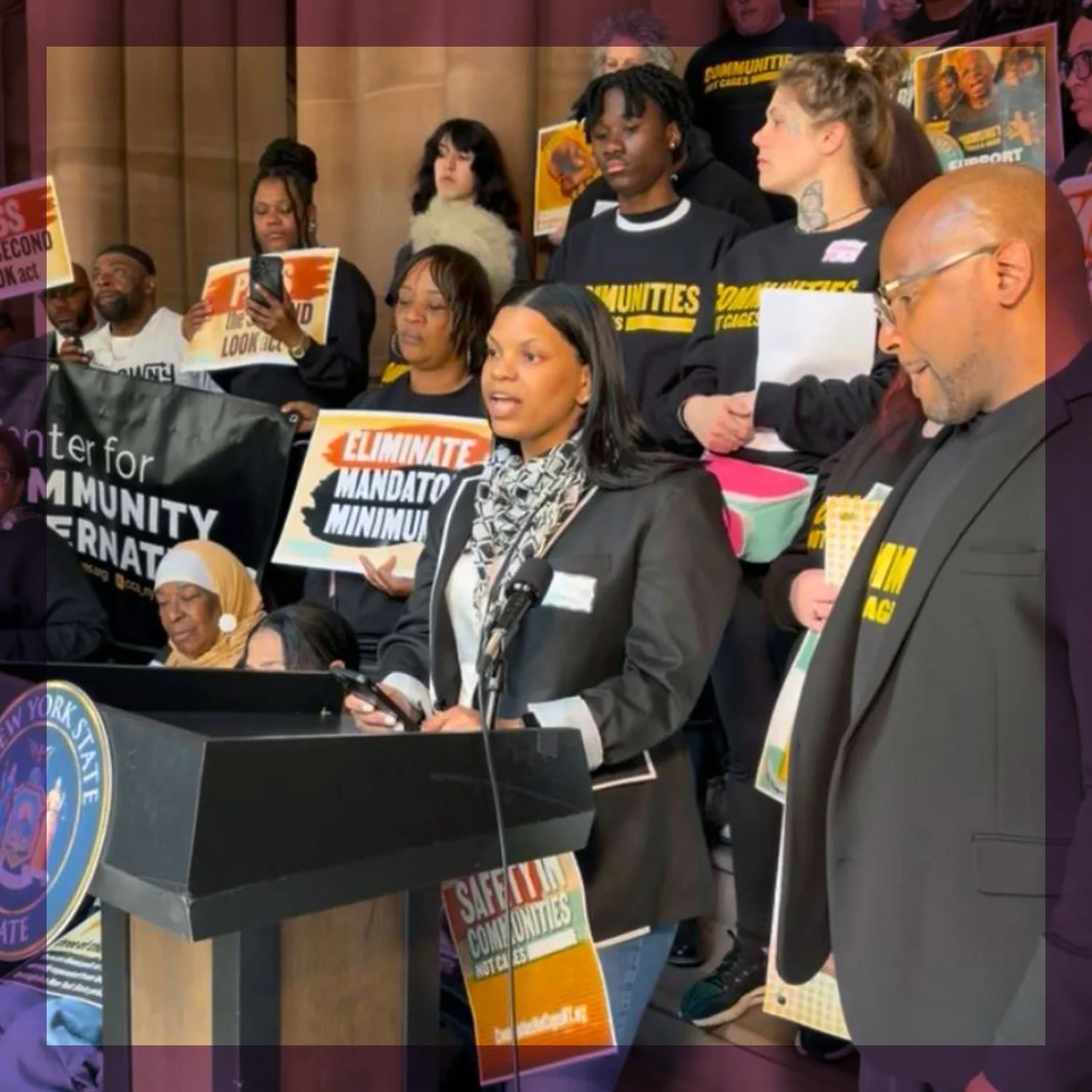 A group of people gathered at a press conference or protest, holding signs and banners that read 'Eliminate Mandatory Minimums' and 'Save Our Communities,' with some individuals speaking at a podium and others listening attentively.