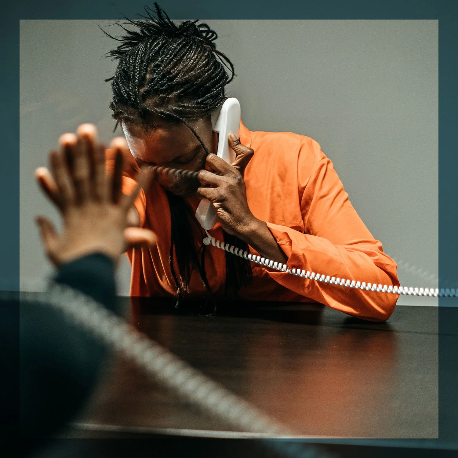 Man with dreadlocks in an orange prison jumpsuit talking on a landline telephone with his eyes closed and hand raised, sitting at a dark table.