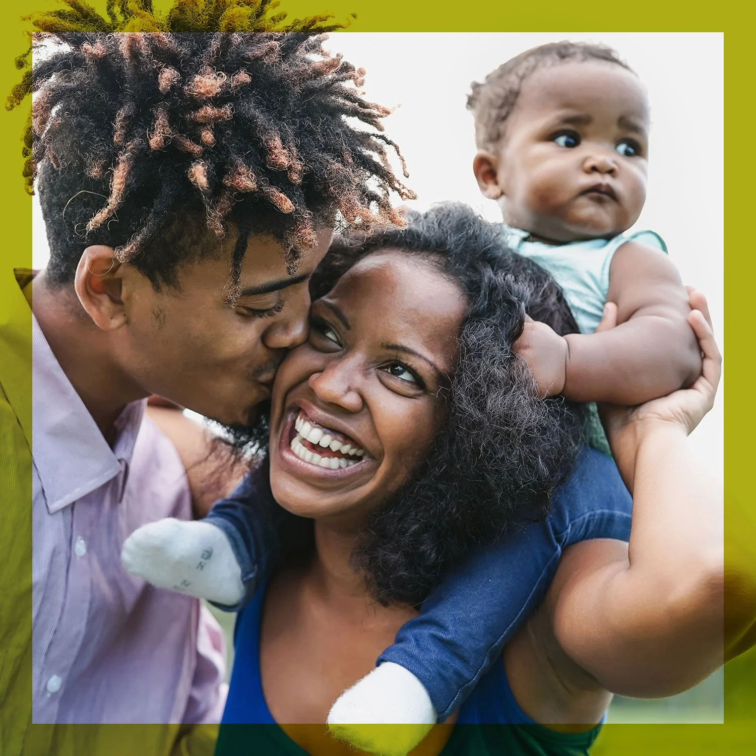 A family celebrating outdoors, with a man kissing a woman on the cheek, both smiling, and a young child held above them.