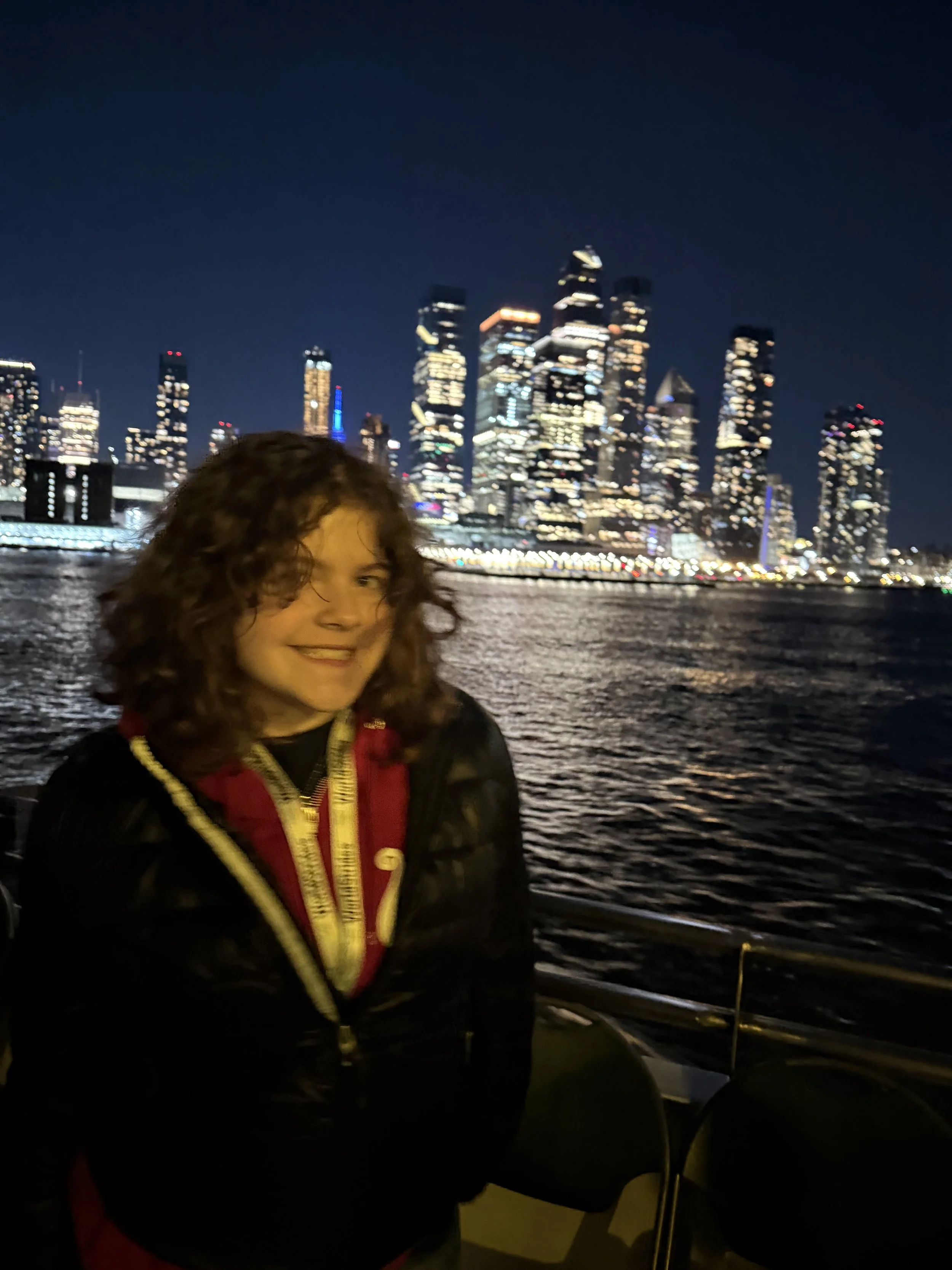 A person with curly brown hair smiling at night with a city skyline lit up in the background across a body of water.