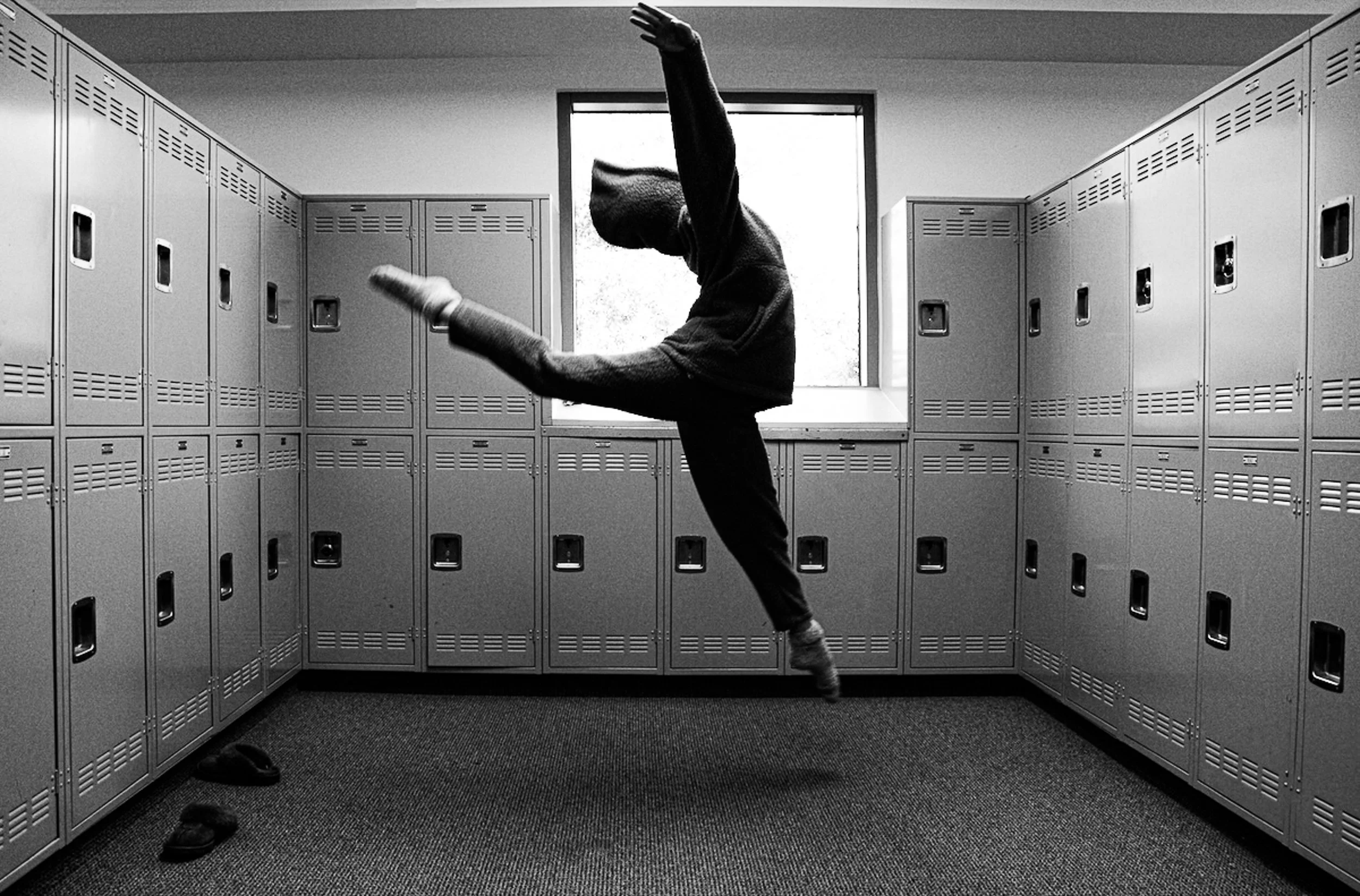 A person wearing a hoodie and gloves performing a high kick inside a locker room, surrounded by gray lockers.