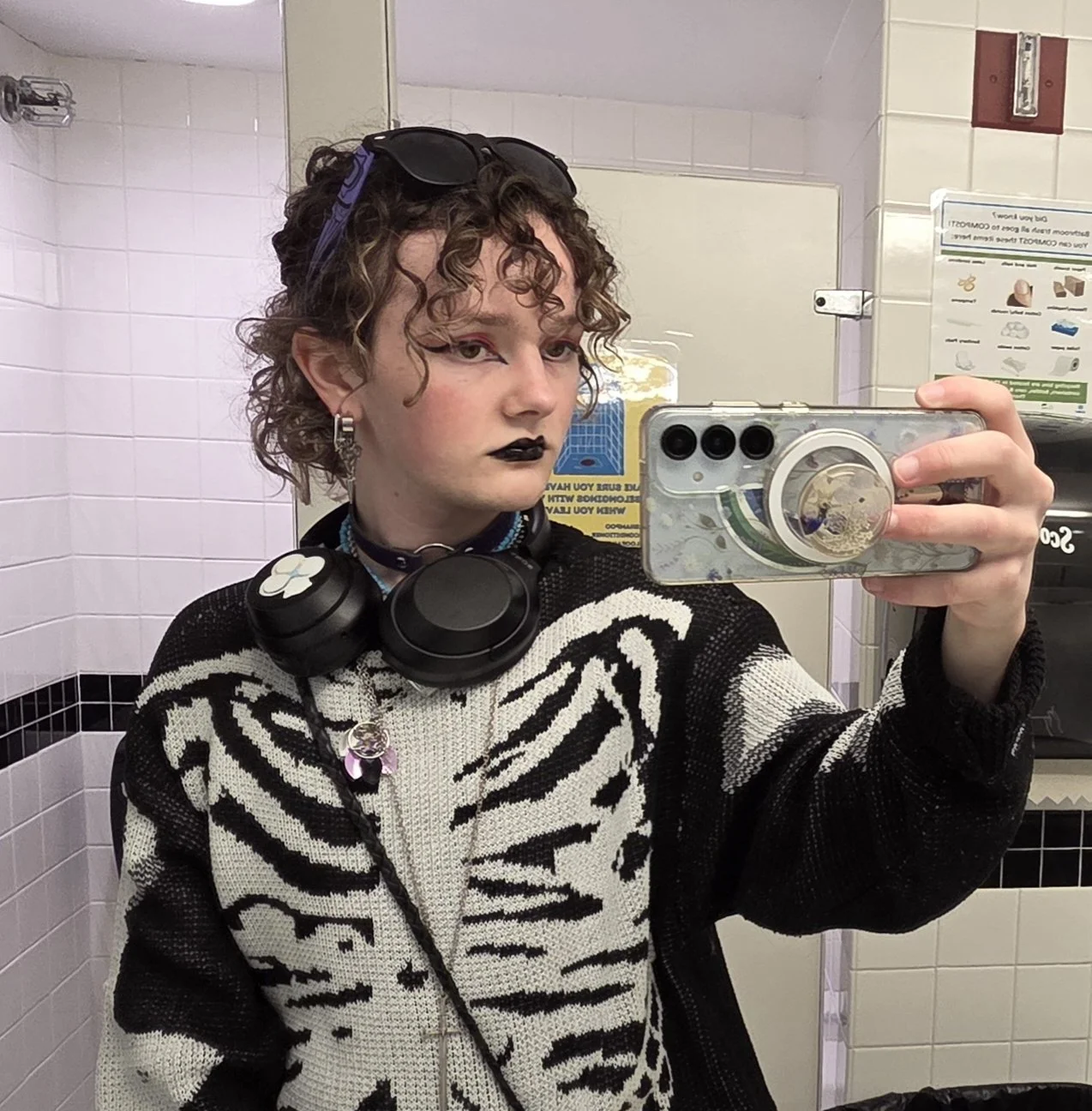 A person with curly hair and makeup taking a mirror selfie in a public restroom. They are wearing a black and white zebra-striped sweater, black lipstick, and have headphones around their neck.