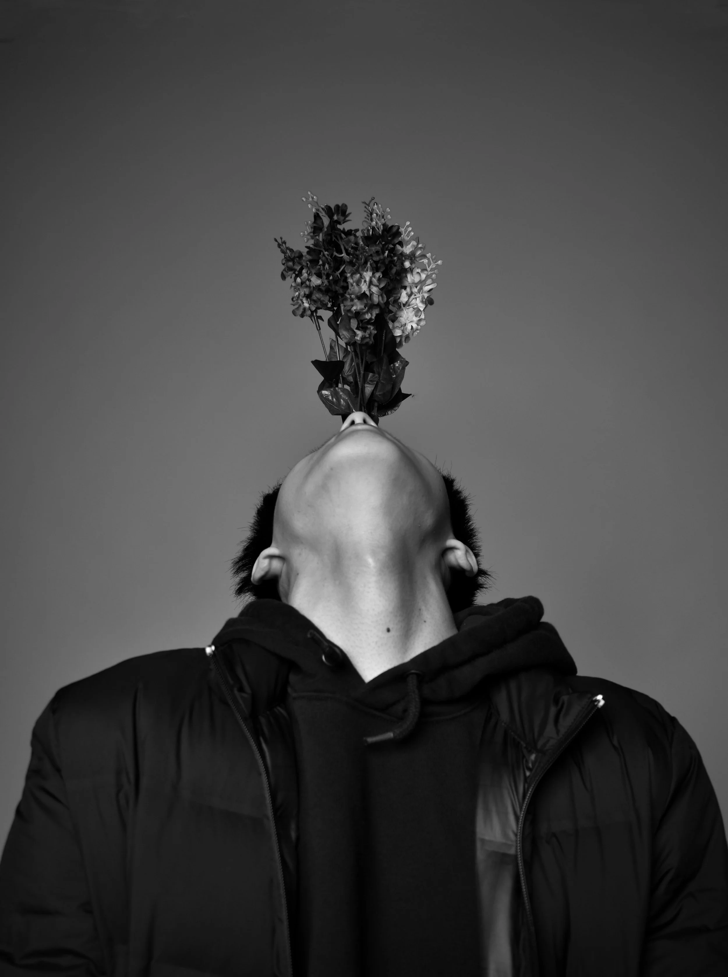 A person with dark hair and light skin, wearing a black jacket, is holding a bouquet of flowers upside down near their face.