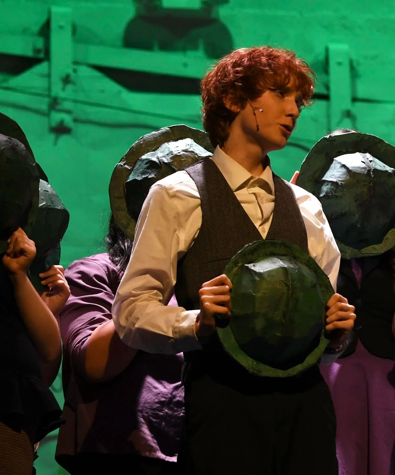 A person with curly red hair wearing a white shirt and dark vest holding a large green leafy object, surrounded by others with similar objects on their heads, against a green background.
