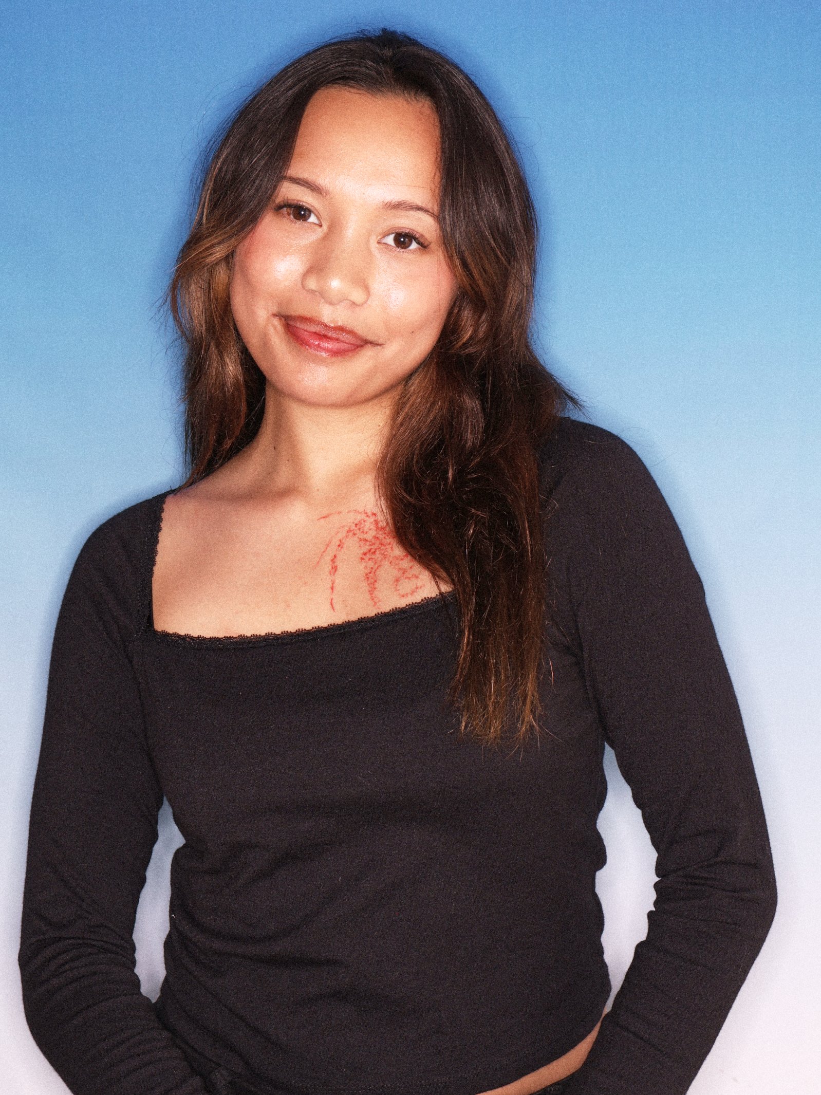 A woman with long brown hair, wearing a black top with a red tattoo on her chest, sitting against a blue background.
