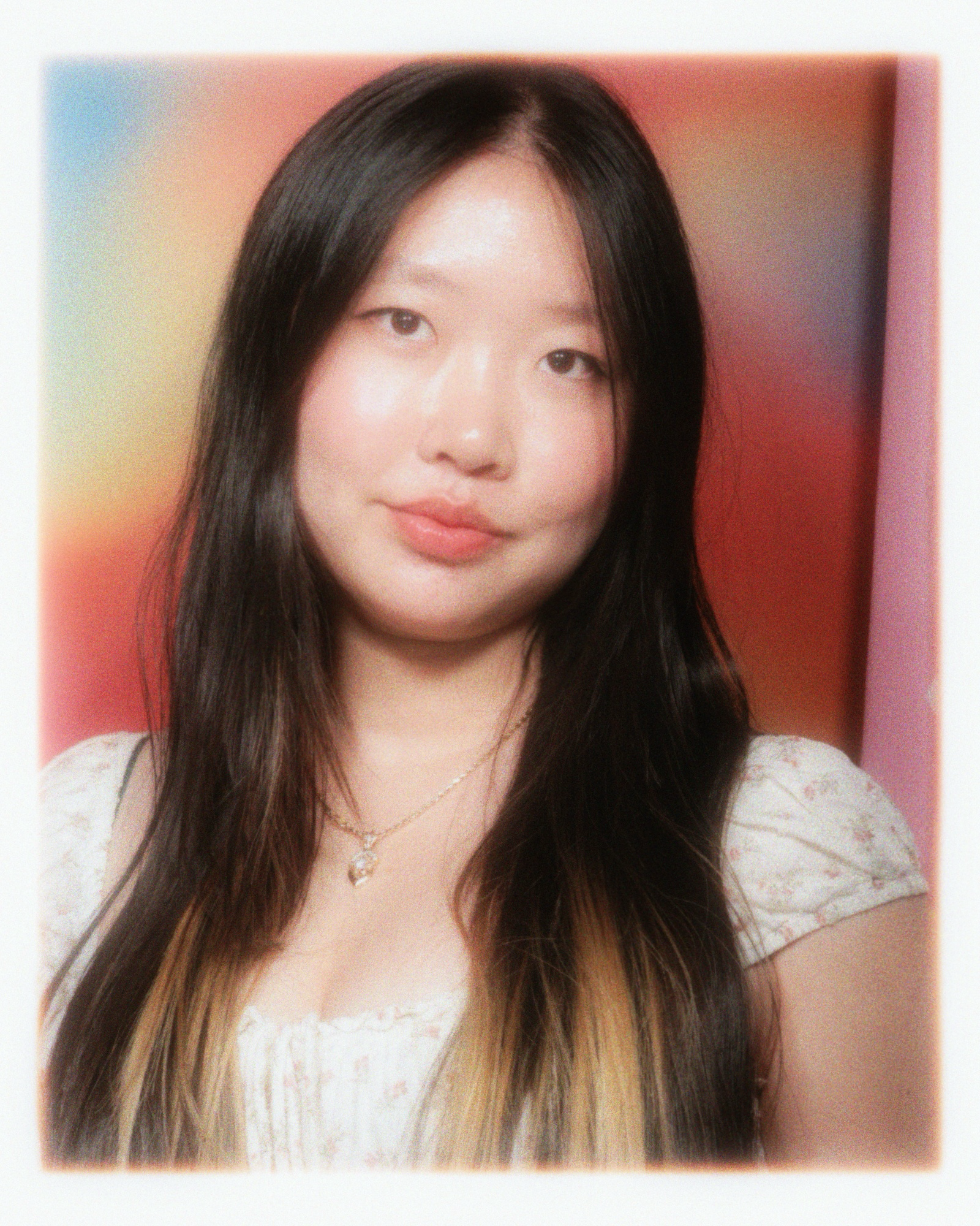 A young woman with long dark hair and light makeup taking a selfie against a pink and beige background, wearing a white lace top and layered necklaces.
