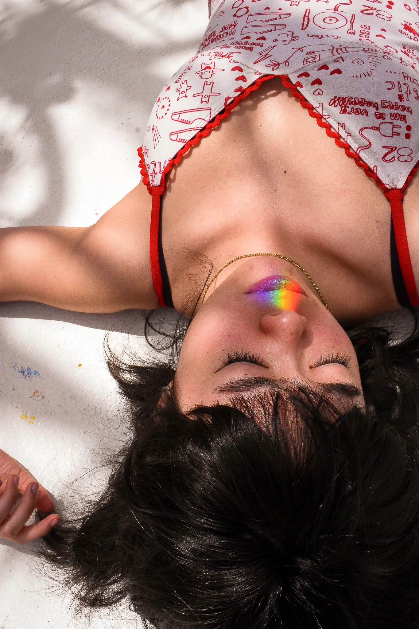 A young woman with dark hair lying on a white textured surface, wearing a white tank top with red drawings and red straps. She has closed eyes and a rainbow reflection on her lips, with her hand resting on the surface near her head.