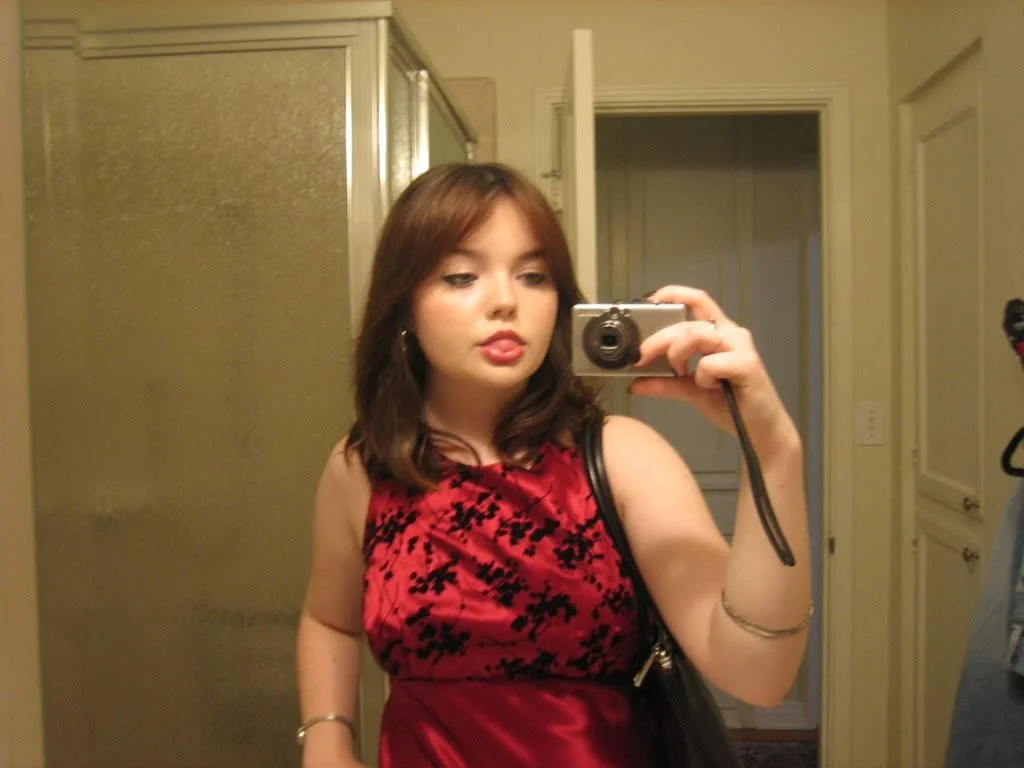 A young woman taking a mirror selfie in a bathroom, wearing a red and black dress, with shoulder-length brown hair.