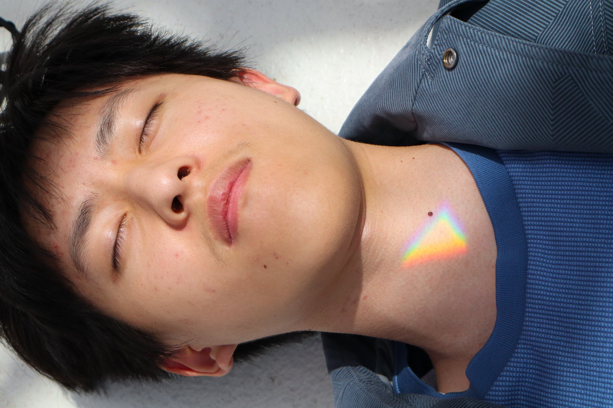 A close-up of a young person's face and upper torso, with their eyes closed, lying down on a light-colored surface. The person has short dark hair and a few acne spots on their face. There is a small rainbow reflection on their neck. They are wearing a blue shirt or jacket.