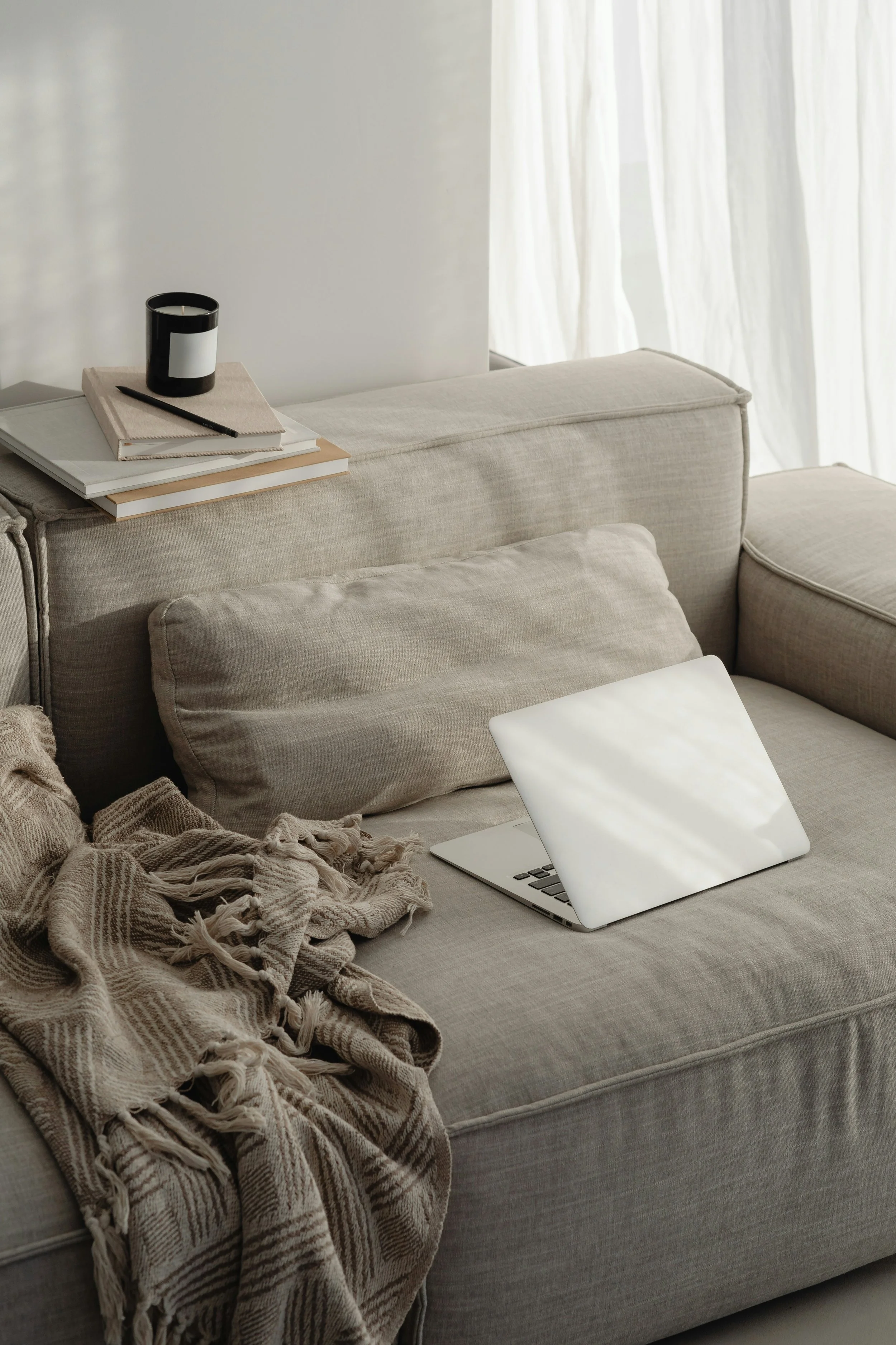 A beige sofa with a laptop, a plaid blanket, and a throw pillow. On the sofa's armrest, there's a stack of books, a notebook, a black pen, and a black candle with a white label. In the background, white curtains cover a window, allowing natural light to filter into the room.