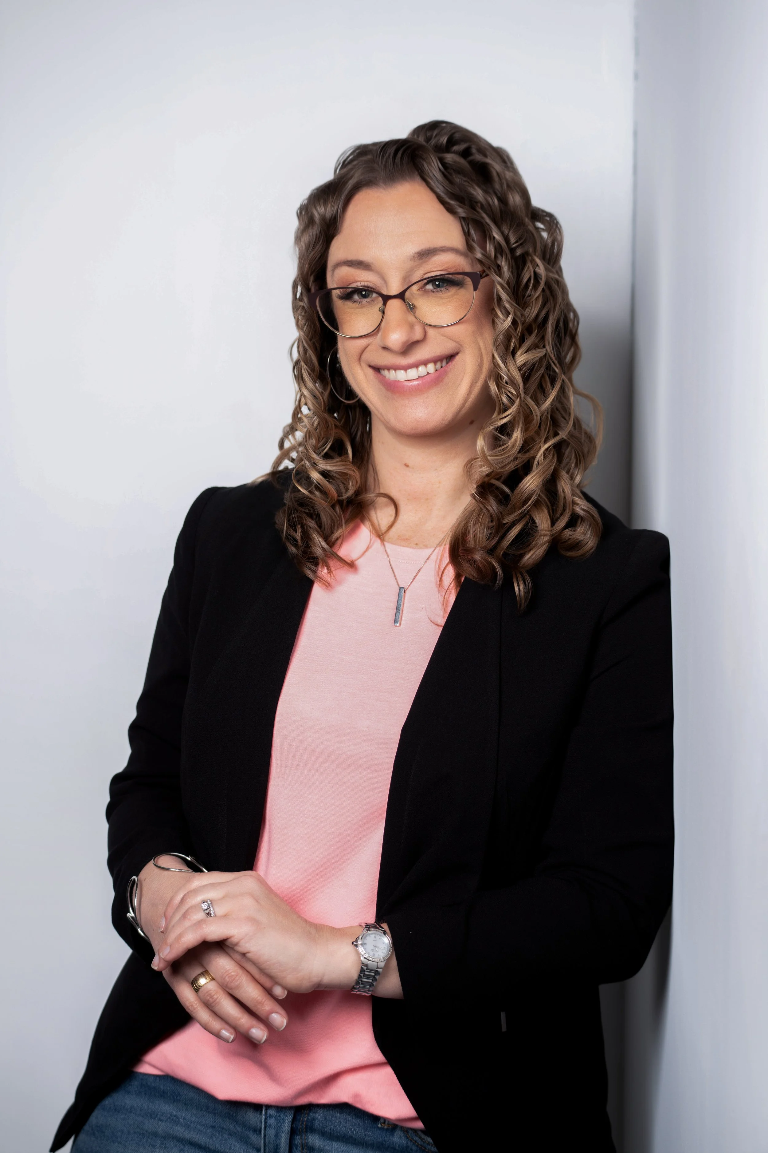 Portrait of a woman with curly brown hair wearing glasses, a black blazer, and a pink top, smiling and crossing her hands in front of her.