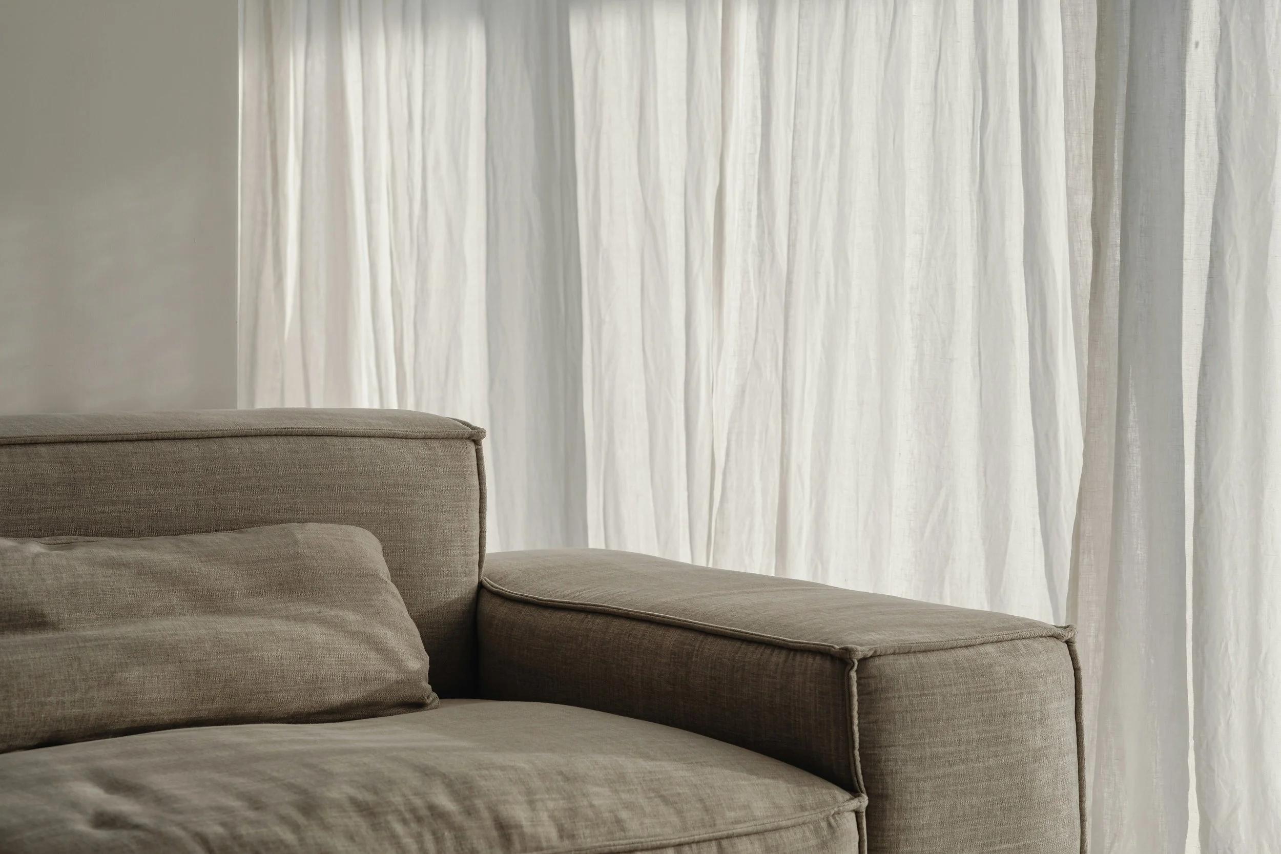 A beige sectional sofa with cushions in front of a white sheer curtain.