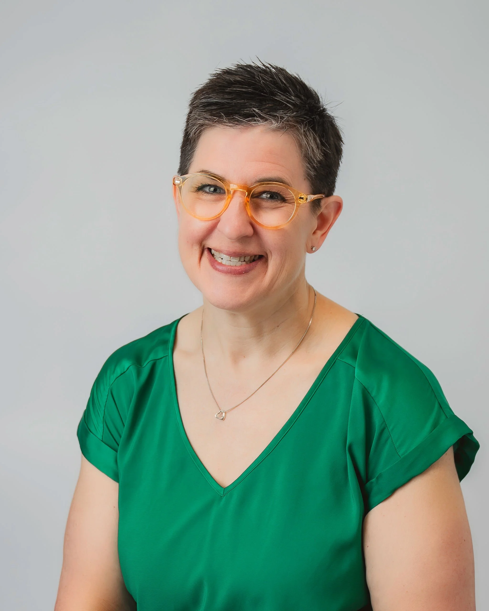 A woman with short dark hair, wearing glasses with a clear orange frame, smiling, dressed in a green V-neck top, against a plain light gray background.