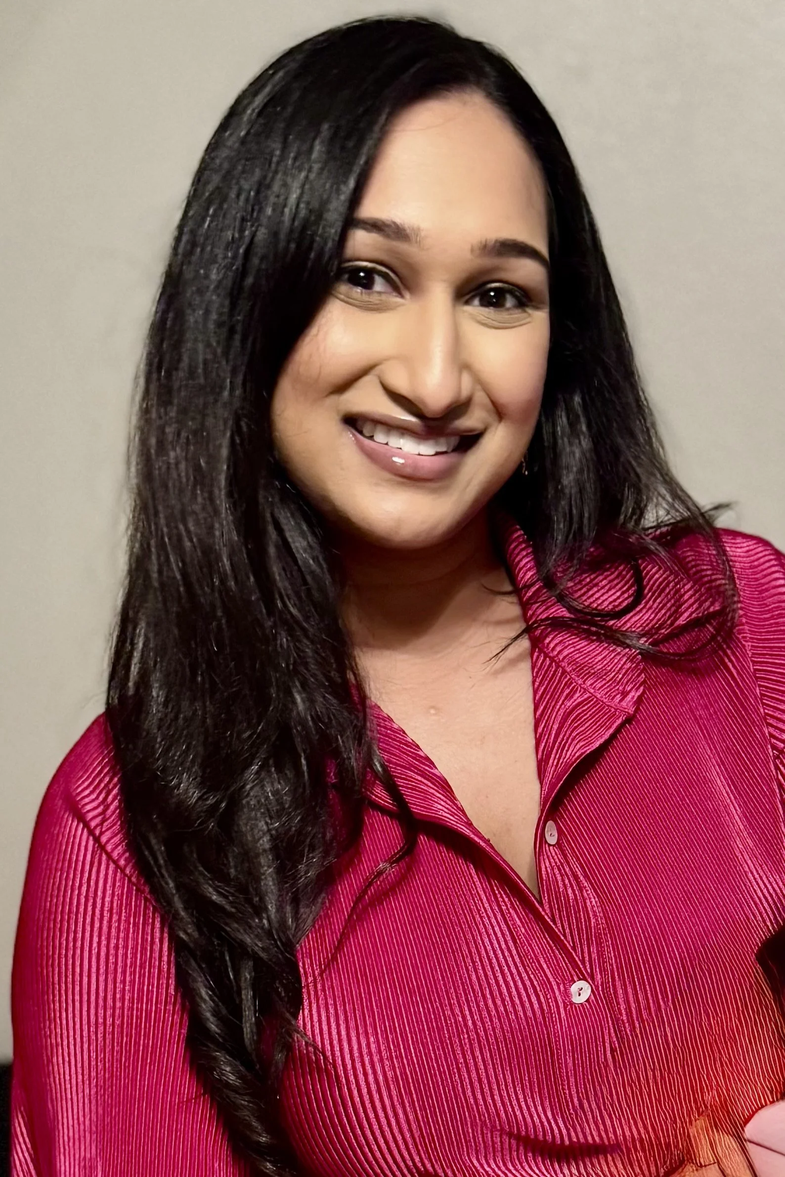 A woman with long black hair wearing a pink, long-sleeved button-up shirt, smiling with her teeth visible.