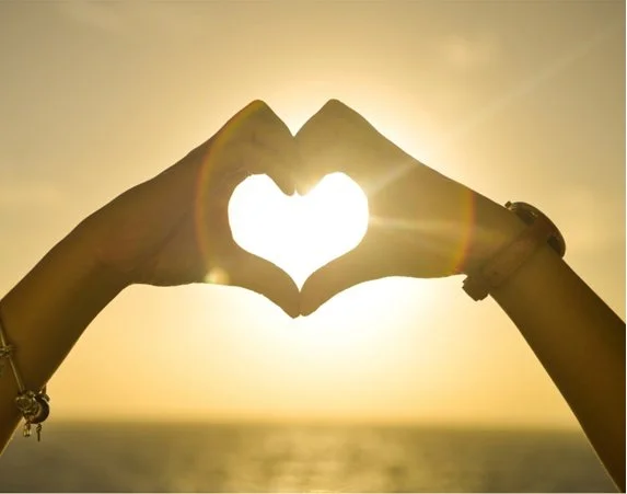 Two hands forming a heart shape with the sun shining through at sunset over the ocean.