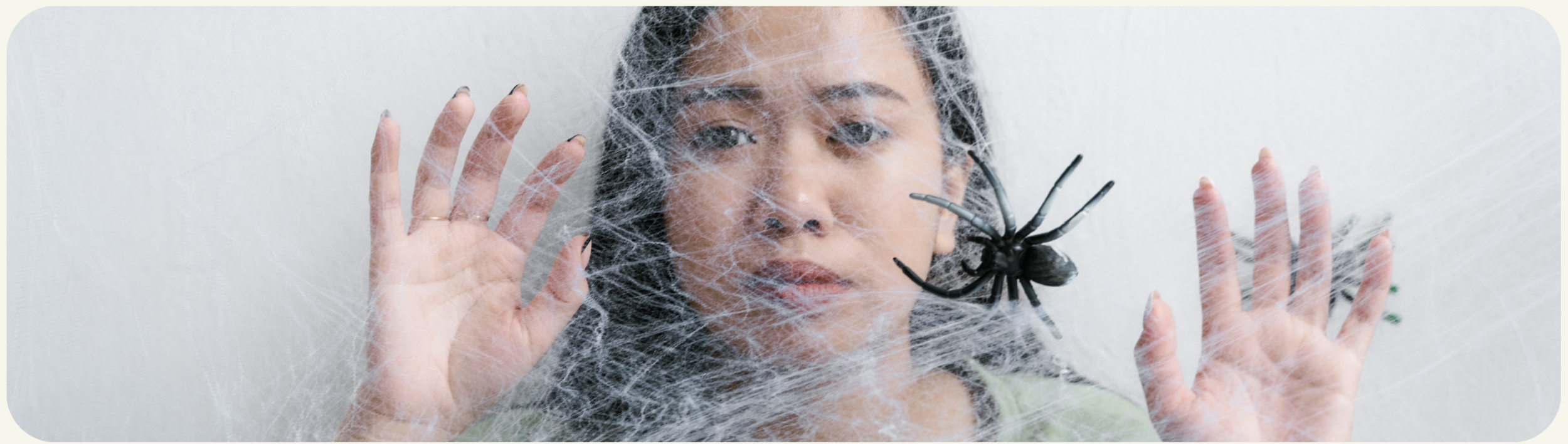 A woman with dark hair looks through a spider web with a large black spider on it, her hands pressed against the web.