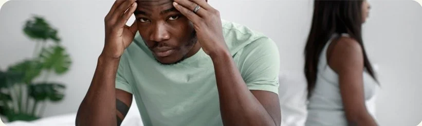 A man sitting on a bed looks distressed, with his hands on his head. A woman is sitting nearby, facing away.