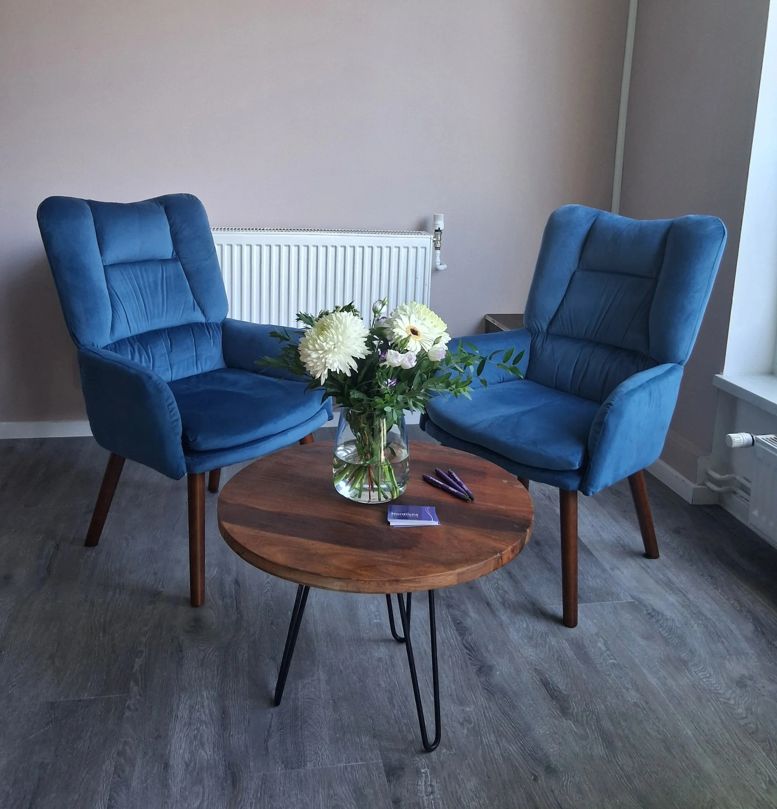 Drei blaue Stühle um einen runden Holzttisch, auf dem ein Blumenstrauß mit weißen Blumen steht, im Raum mit grauem Boden und hellgrauem Wand.