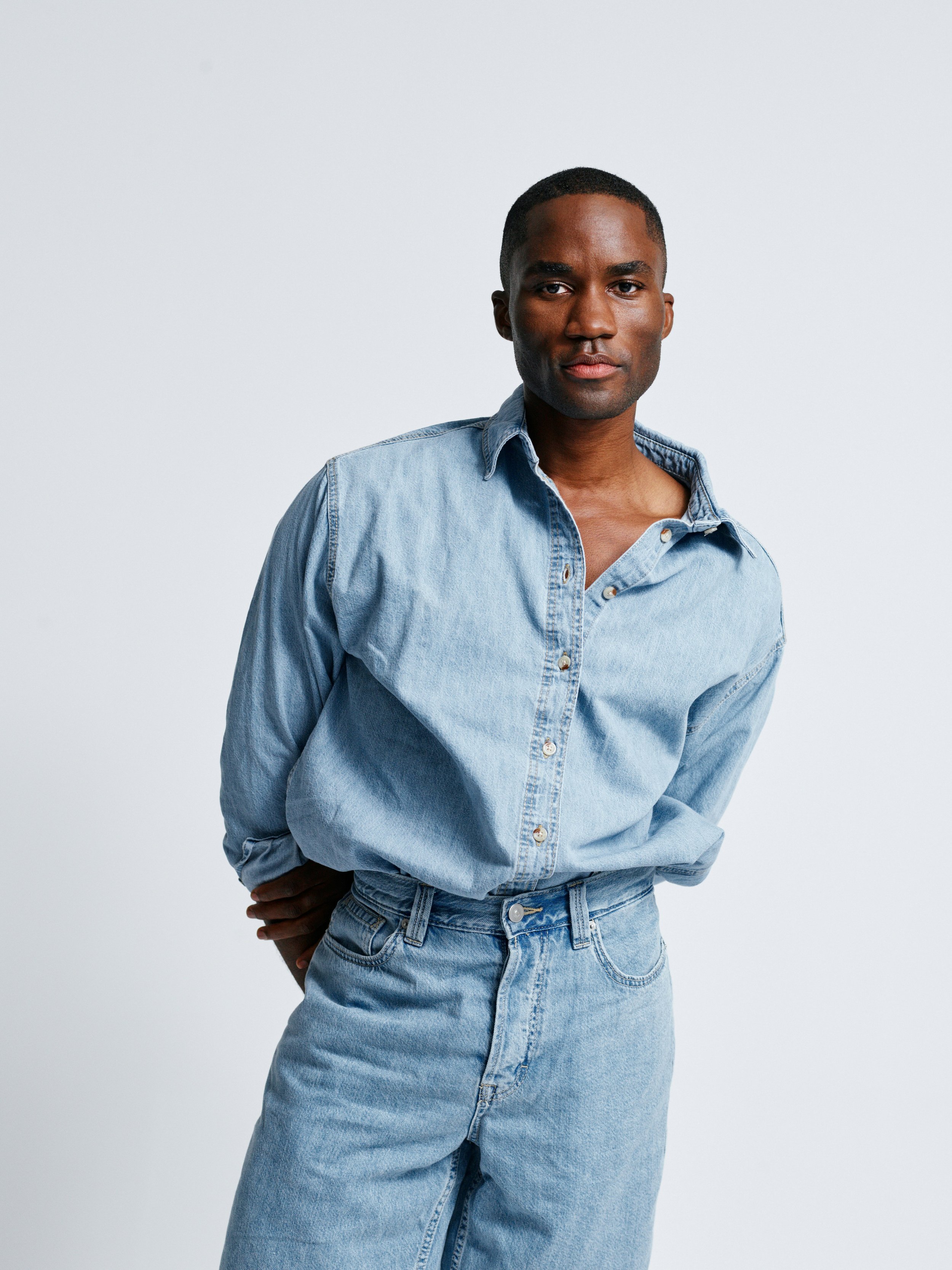 A person with dark skin and short black hair, wearing a light blue denim shirt and jeans, standing against a plain white background.
