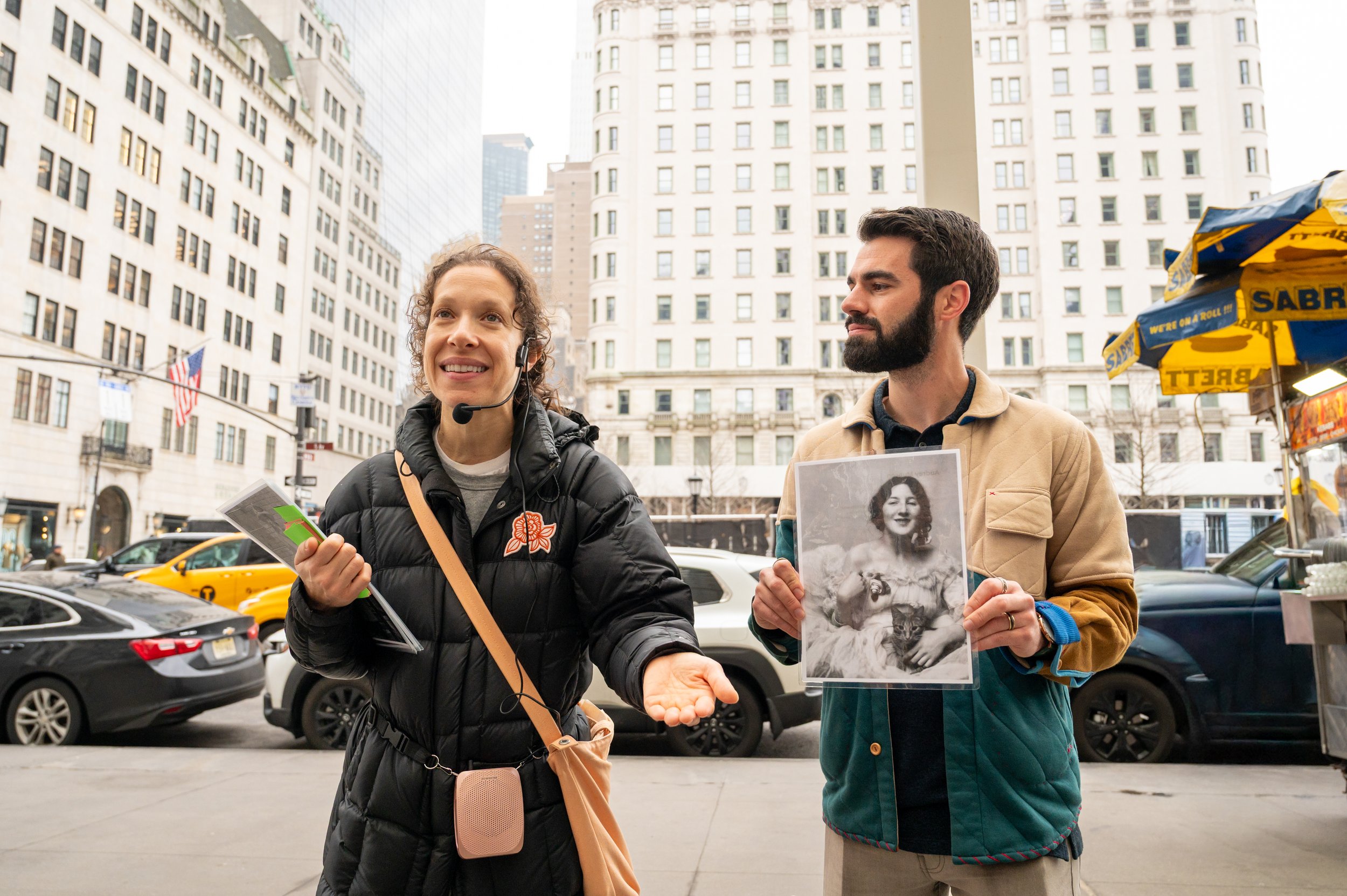 A tour guide wearing a headset and wearing a black puffer jacket talks while a young male guest with a beard stands next to her holding a portrait of Audrey Munson.