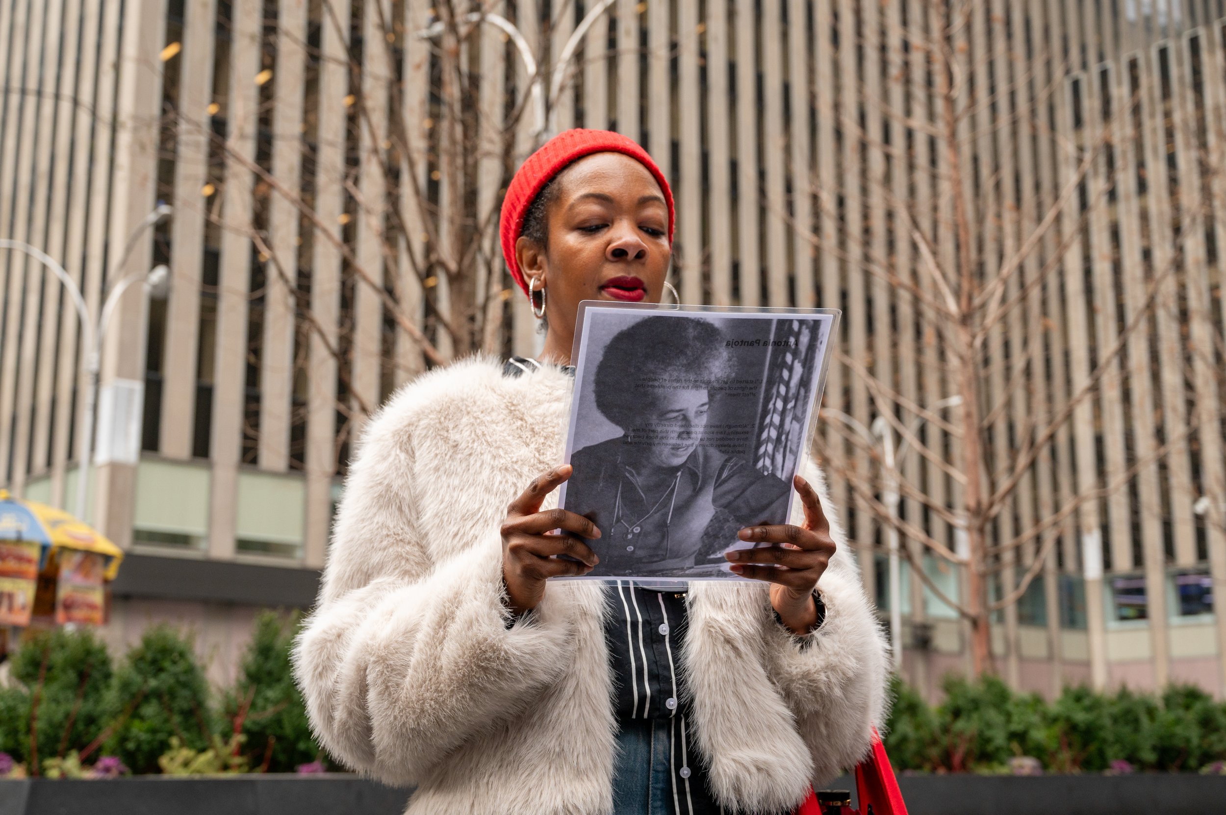 A woman with a red beanie, hoop earrings, and a white fur coat is reading a quote from Antonia Pantoja on the back of her portrait. There is a modern building with vertical metal strips and a leafless tree in the background.