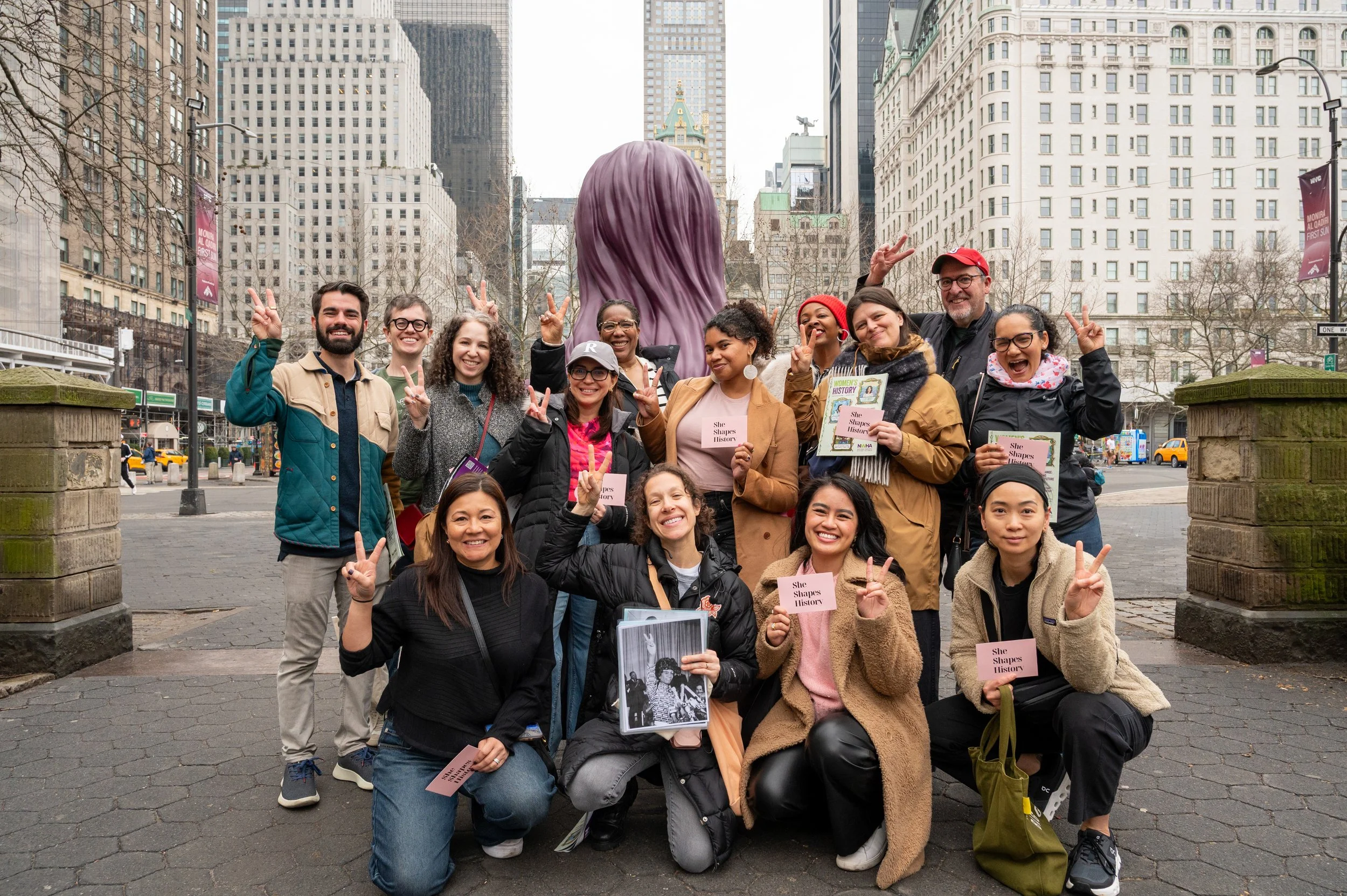 A diverse tour group holding portraits of the women featured on the She Was Here NYC feminist history tour. They are smiling, making peace signs, and the cityscape is visible in the background.