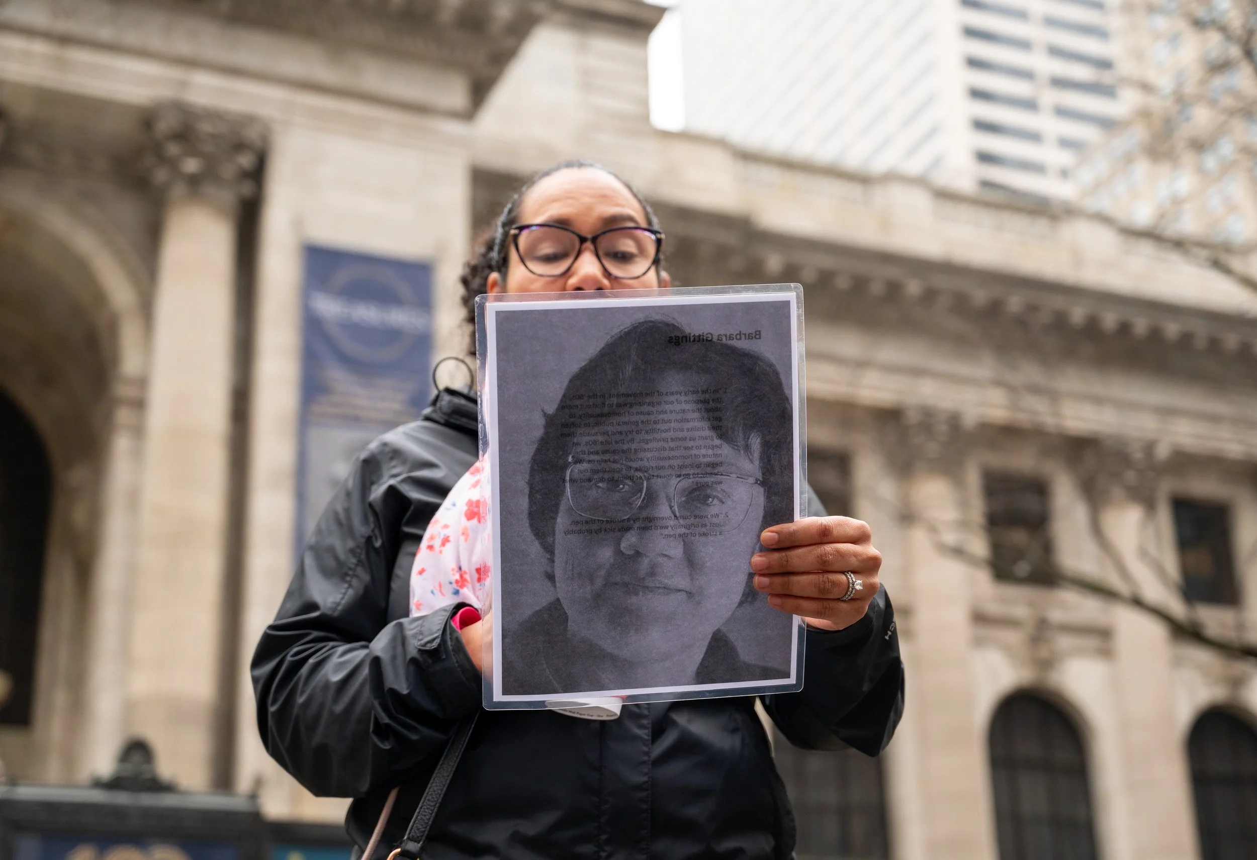 A tour guest wearing glasses and a black jacket holds a printed photograph of a Barbara Gittings face in front of her, in front of the main New York Public Library.
