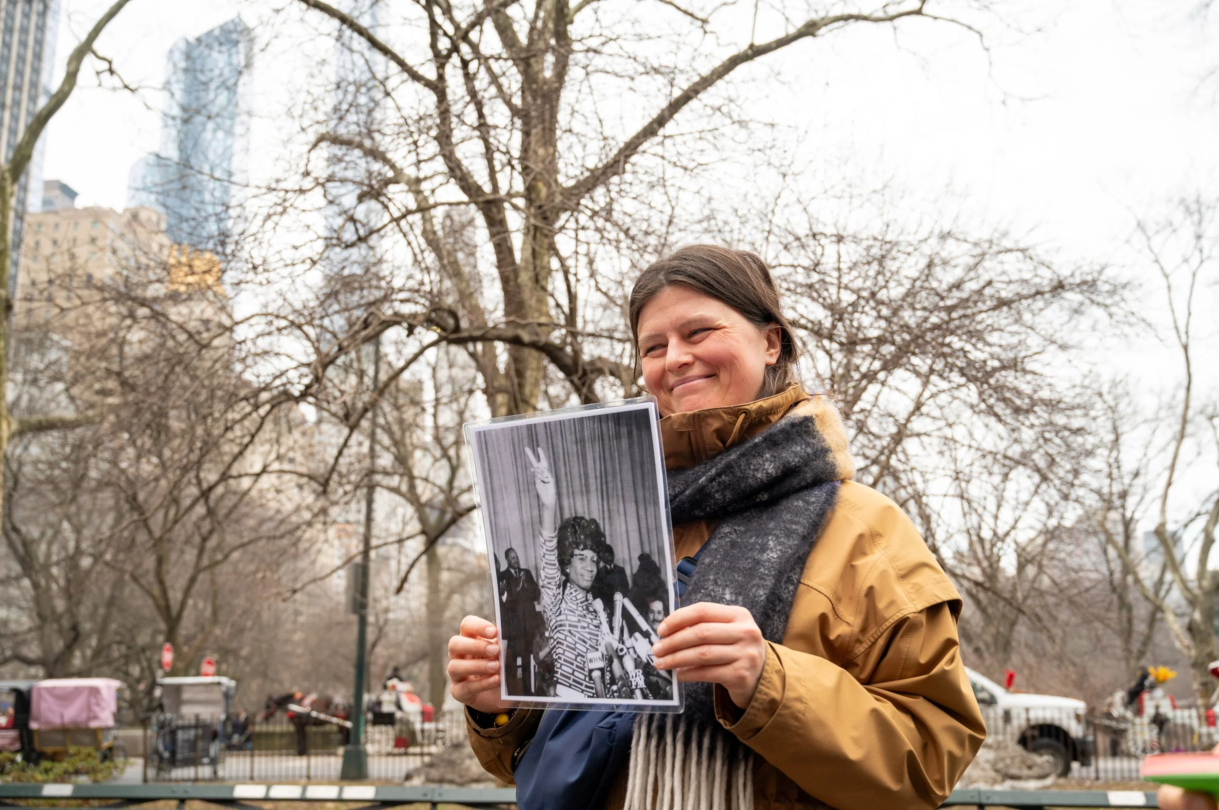A tour guest smiling and holding a photo of Shirley Chisholm in Central Park with leafless trees and city buildings in the background.