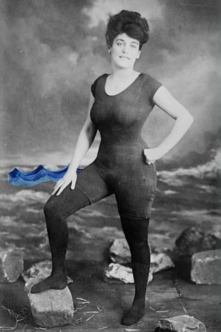 A black-and-white photo of a Annette Kellerman with short, dark hair, wearing a fitted, short-sleeved black outfit, standing outdoors in front of a painted backdrop of rocks and sky. She has one hand on her hip and the other resting on a rock.